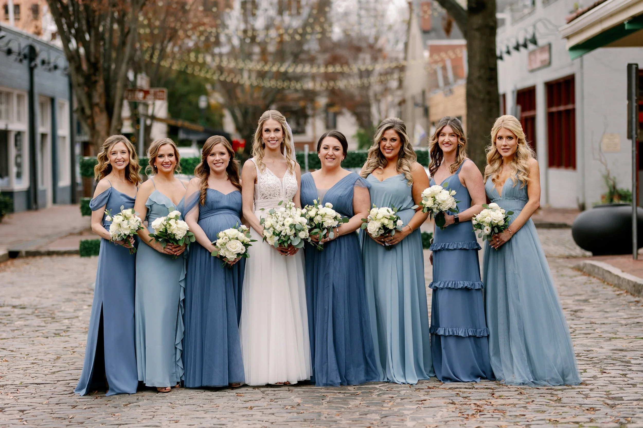 Bride standing on the cobblestone street in City Market Raleigh with her bridesmaids in blue dresses, each holding white and green bouquets during a Market Hall wedding.