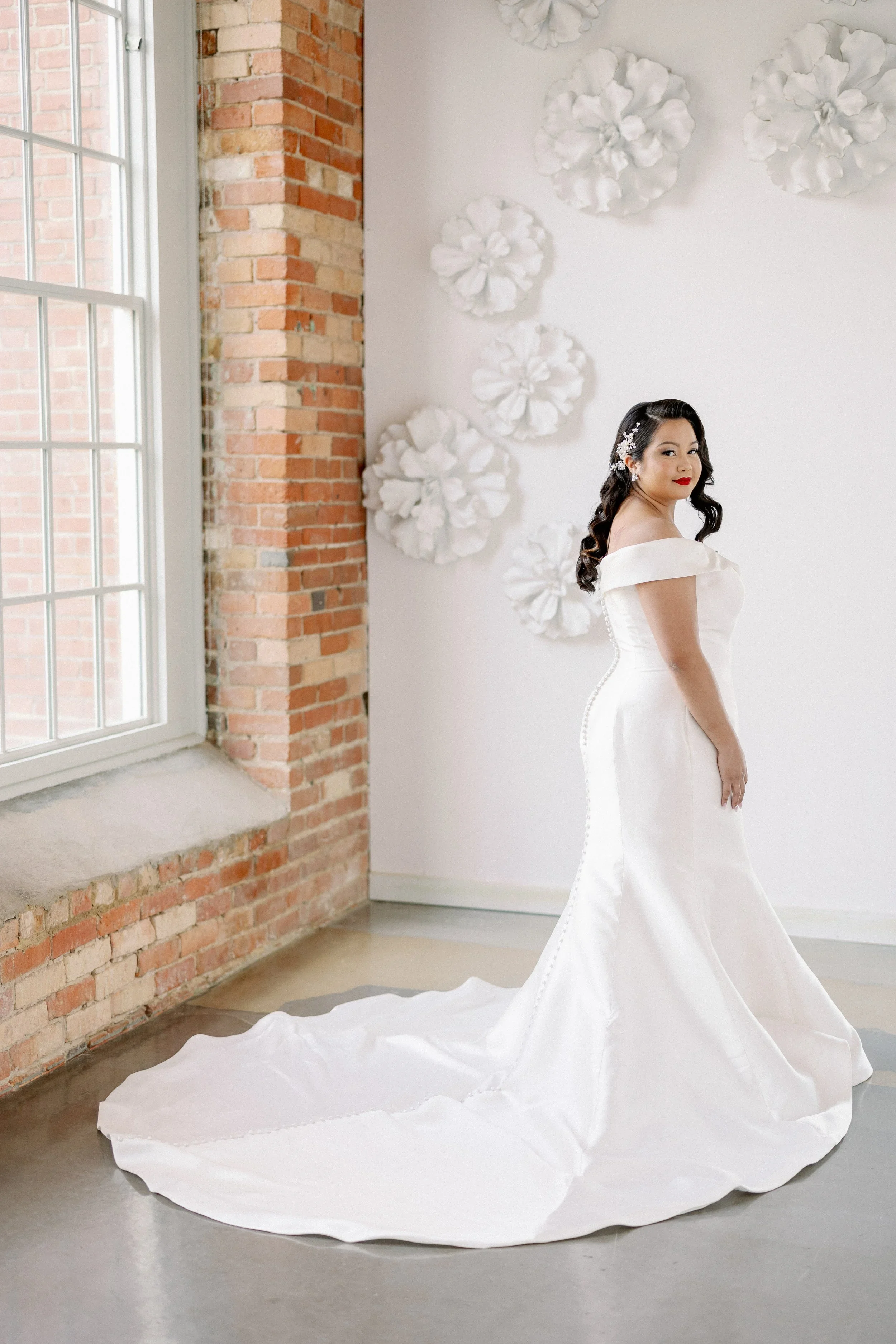 Bride in an off-the-shoulder wedding gown with a long train standing near a large window and exposed brick wall at The Cotton Room in Durham, North Carolina.