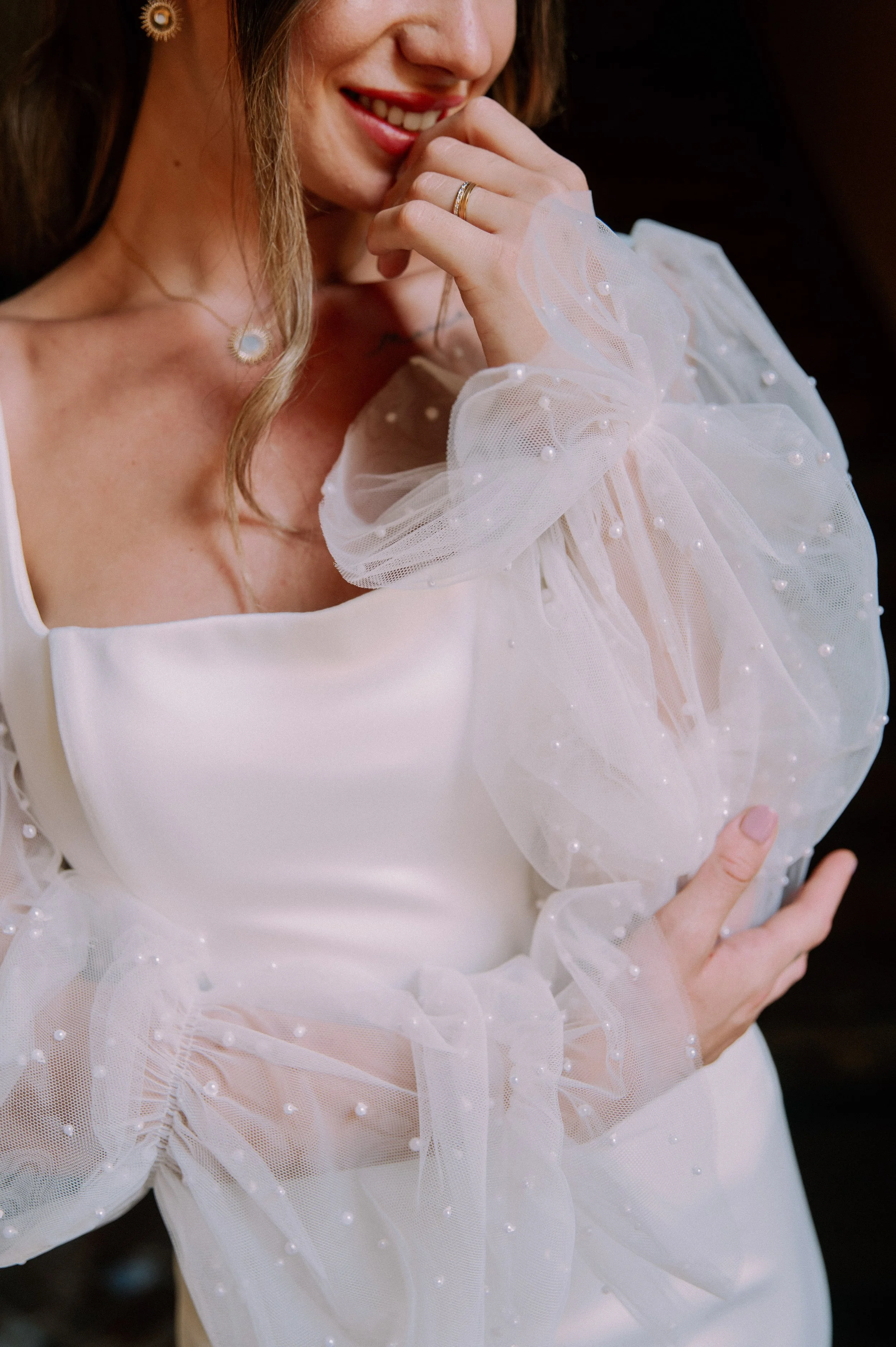 A woman smiling in a white dress with pearl-embellished tulle sleeves, wearing jewelry including a necklace and rings, with her hand near her face.