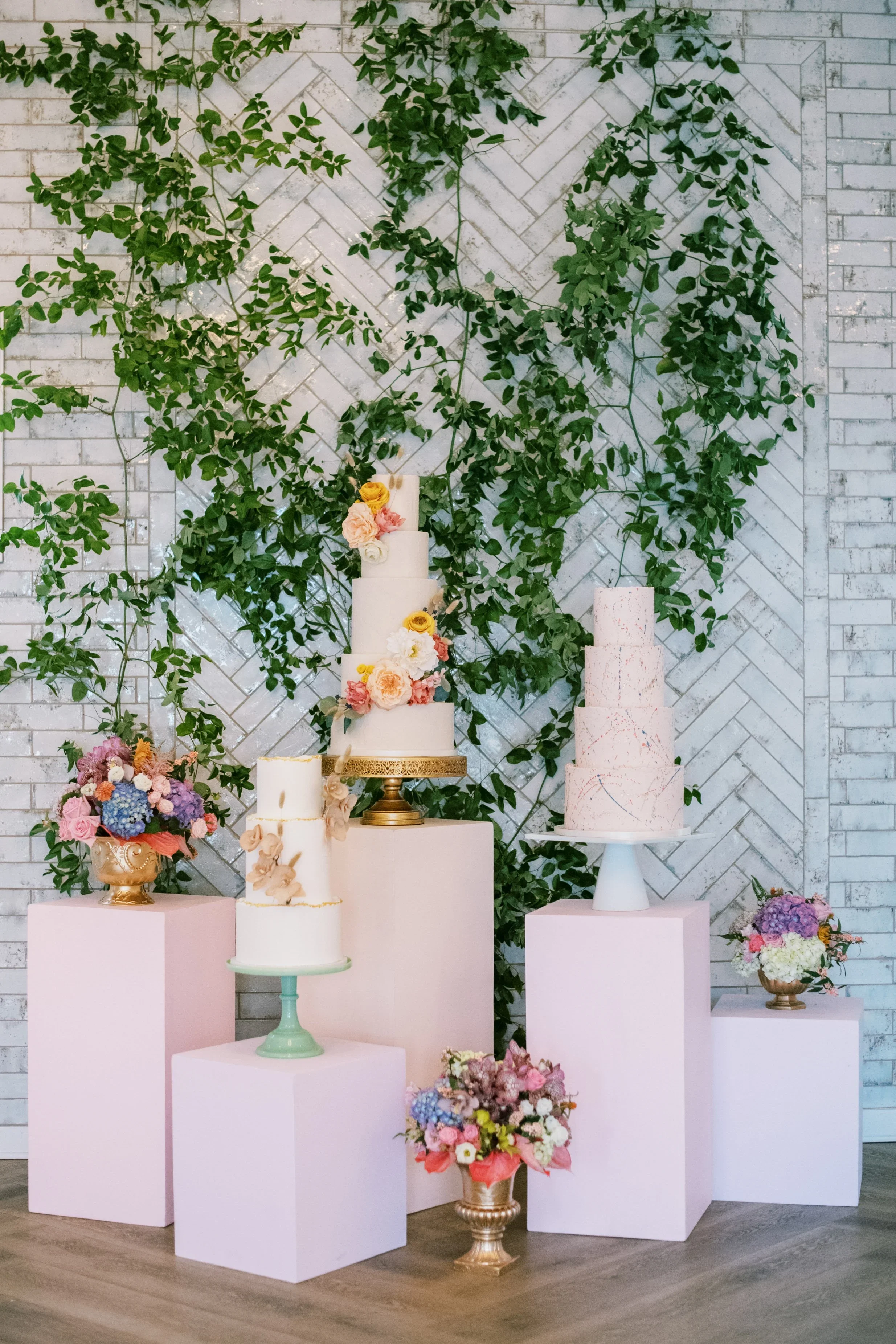 Wedding cake display at The Bradford featuring multiple tiered cakes styled on blush pedestals with pastel florals and greenery against a white brick backdrop.