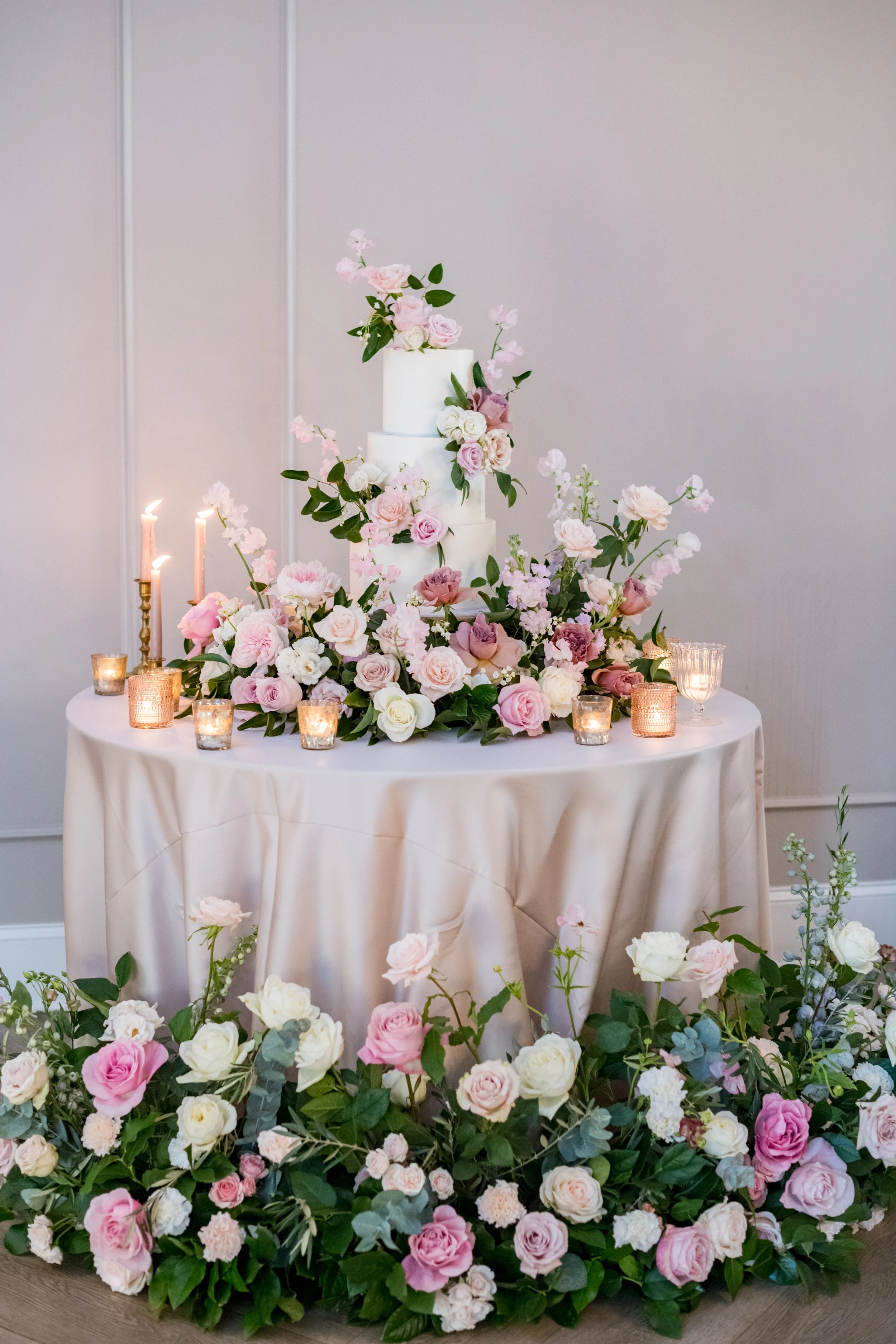 Elegant wedding cake decorated with pink and white flowers, surrounded by candles and lush floral arrangements in soft pastel colors.