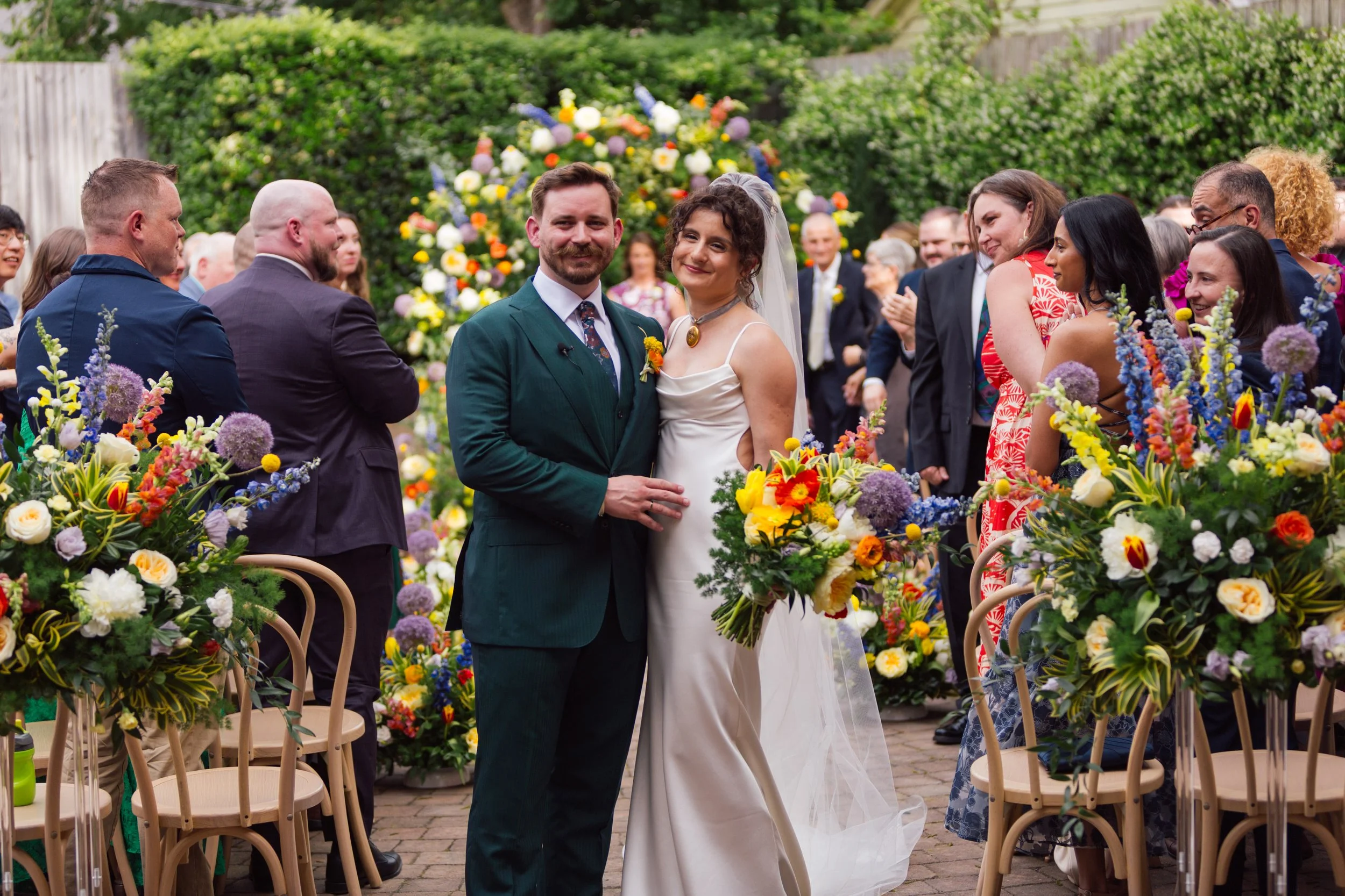 Bride and groom standing together during their outdoor wedding ceremony at The Cookery in Durham, North Carolina, surrounded by guests and vibrant seasonal floral arrangements in the garden courtyard.