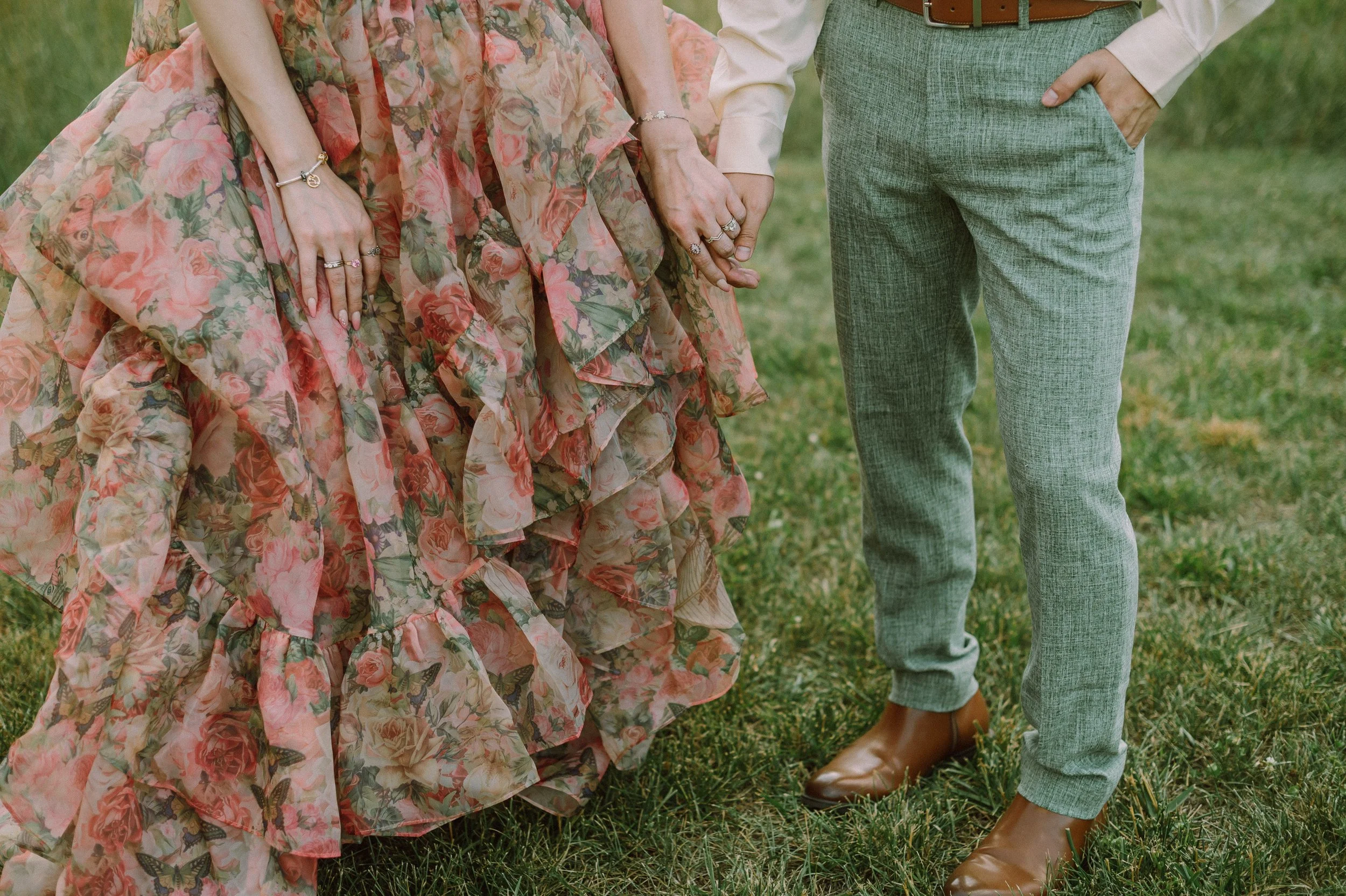 Close-up of a bride's ruffled pink floral wedding dress and stacked rings as she holds hands with her groom in sage green pants at a North Carolina wedding.