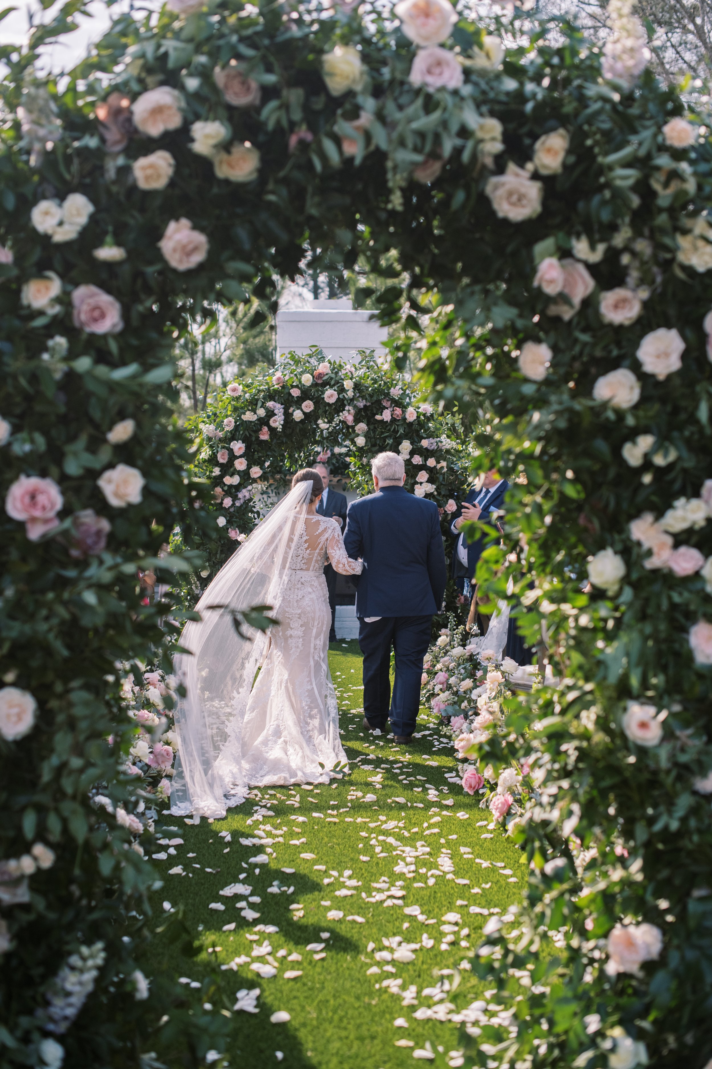 Bride walking down the aisle at The Bradford wedding venue under lush floral arches with rose petals scattered along the grass.