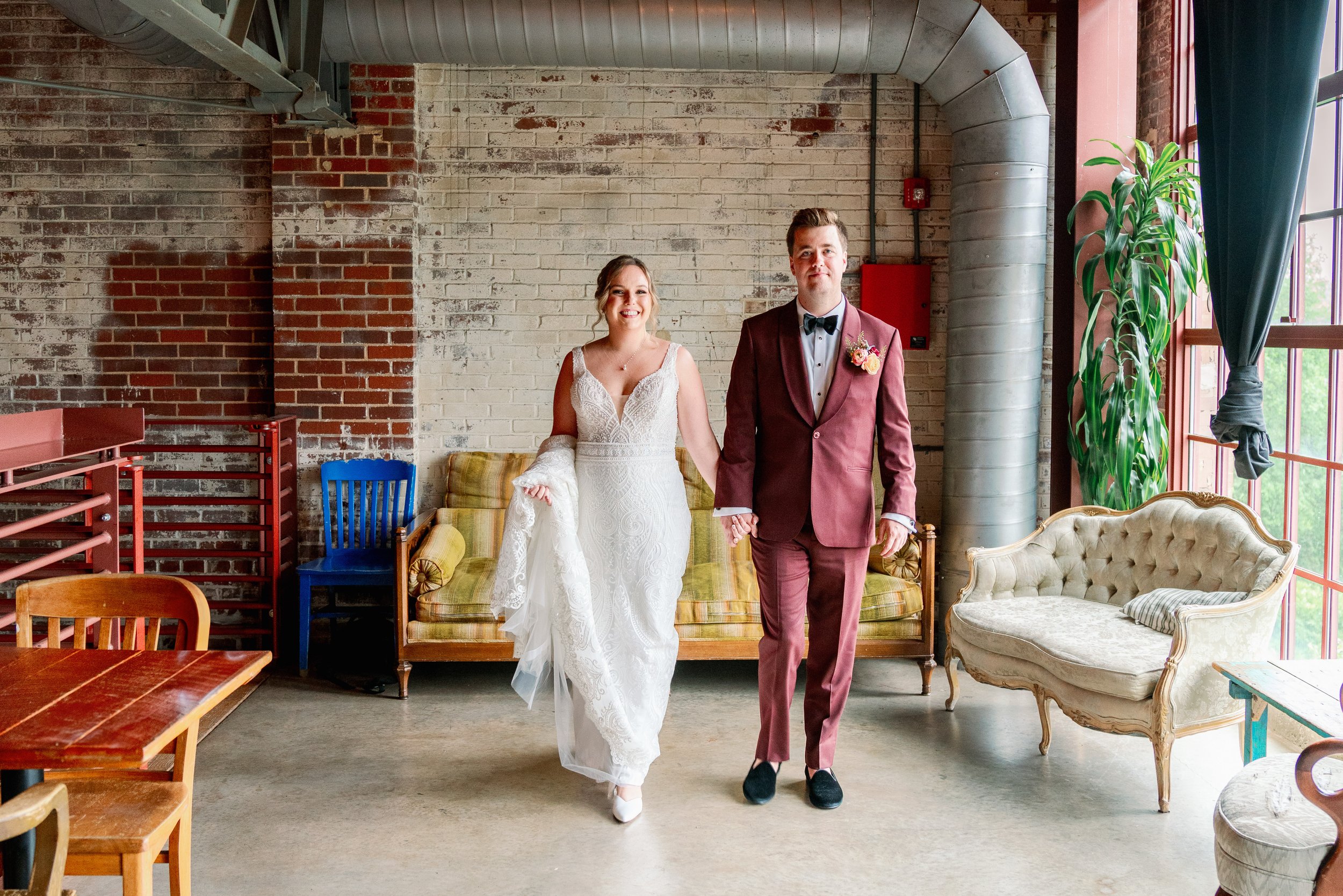 Bride and groom holding hands inside Haw River Ballroom in Saxapahaw North Carolina, photographed in front of exposed brick, vintage furniture, and large industrial windows during their wedding day portraits