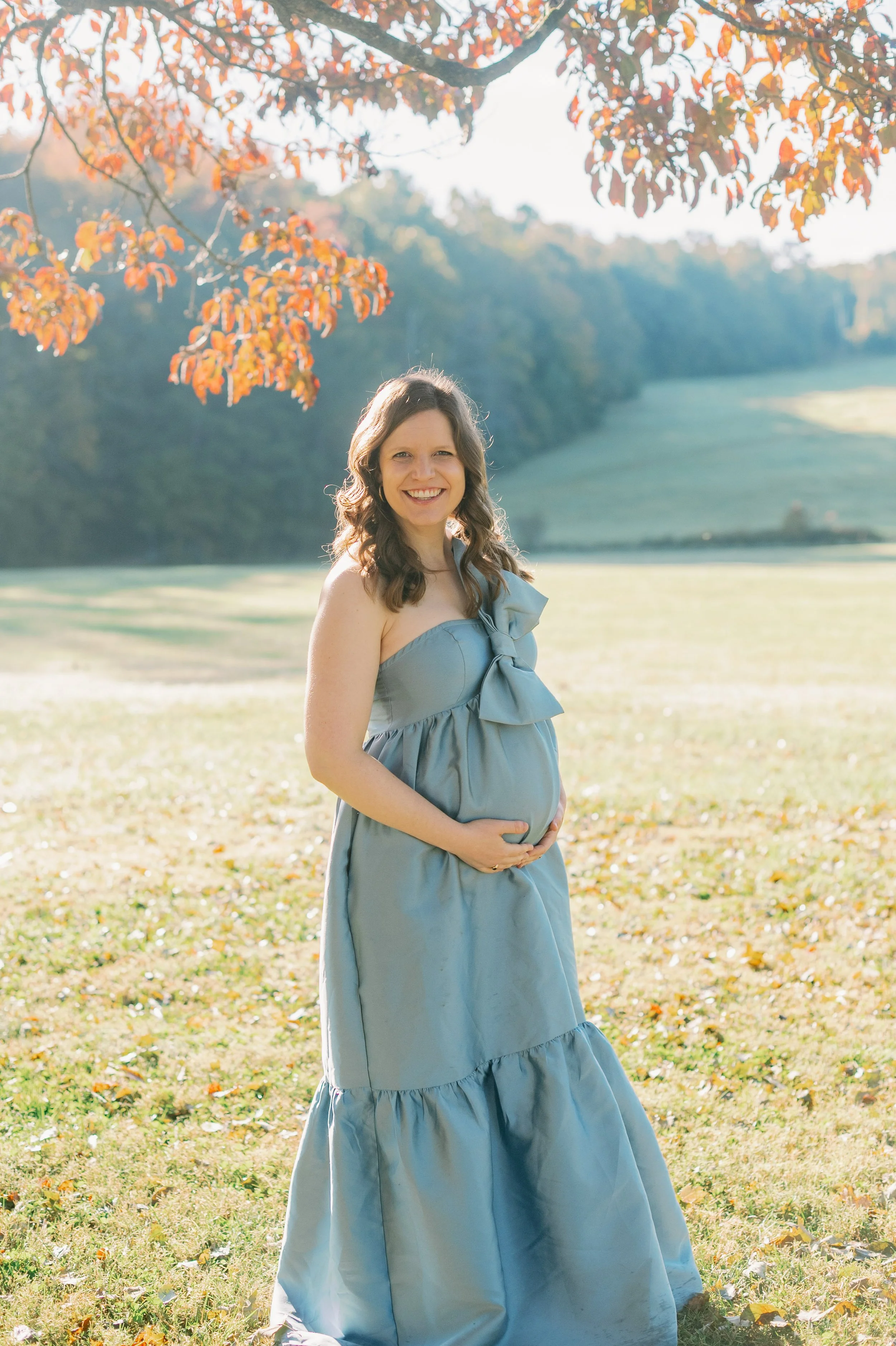 A pregnant woman wearing a sleeveless blue dress with a bow on the chest, standing outdoors in a grassy field with autumn trees and hills in the background.