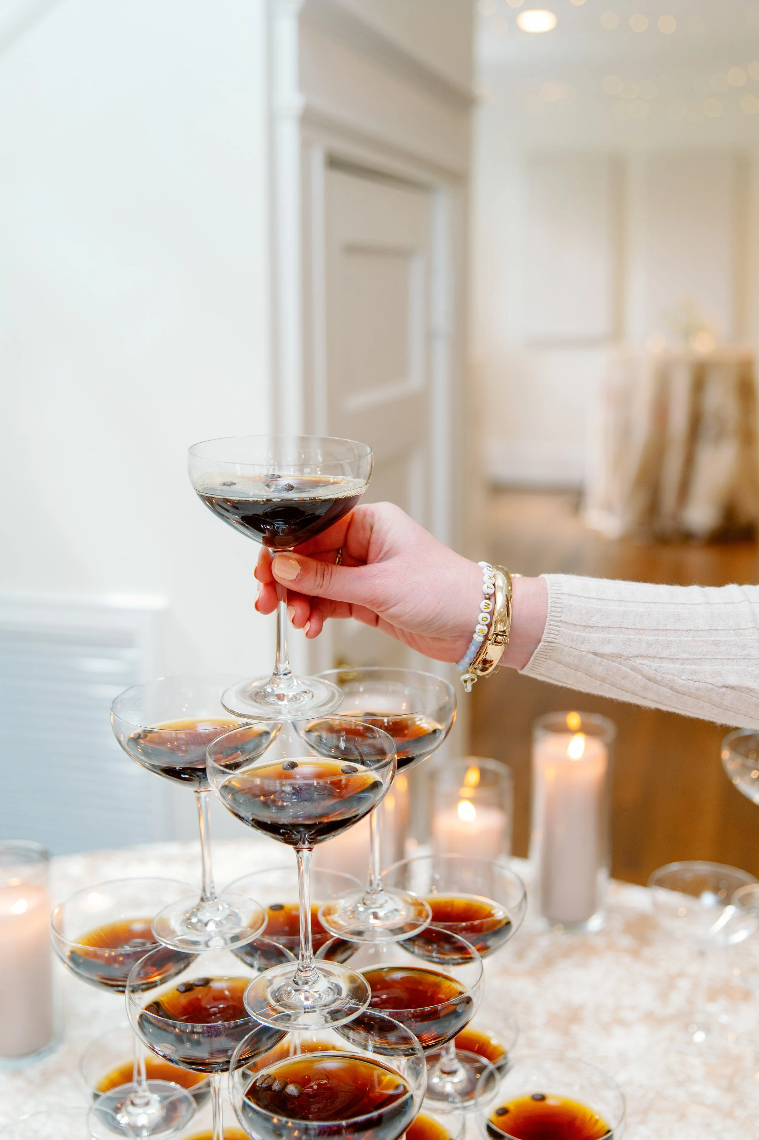 A person stacking glasses filled with dark liquid on a table decorated with lit candles.