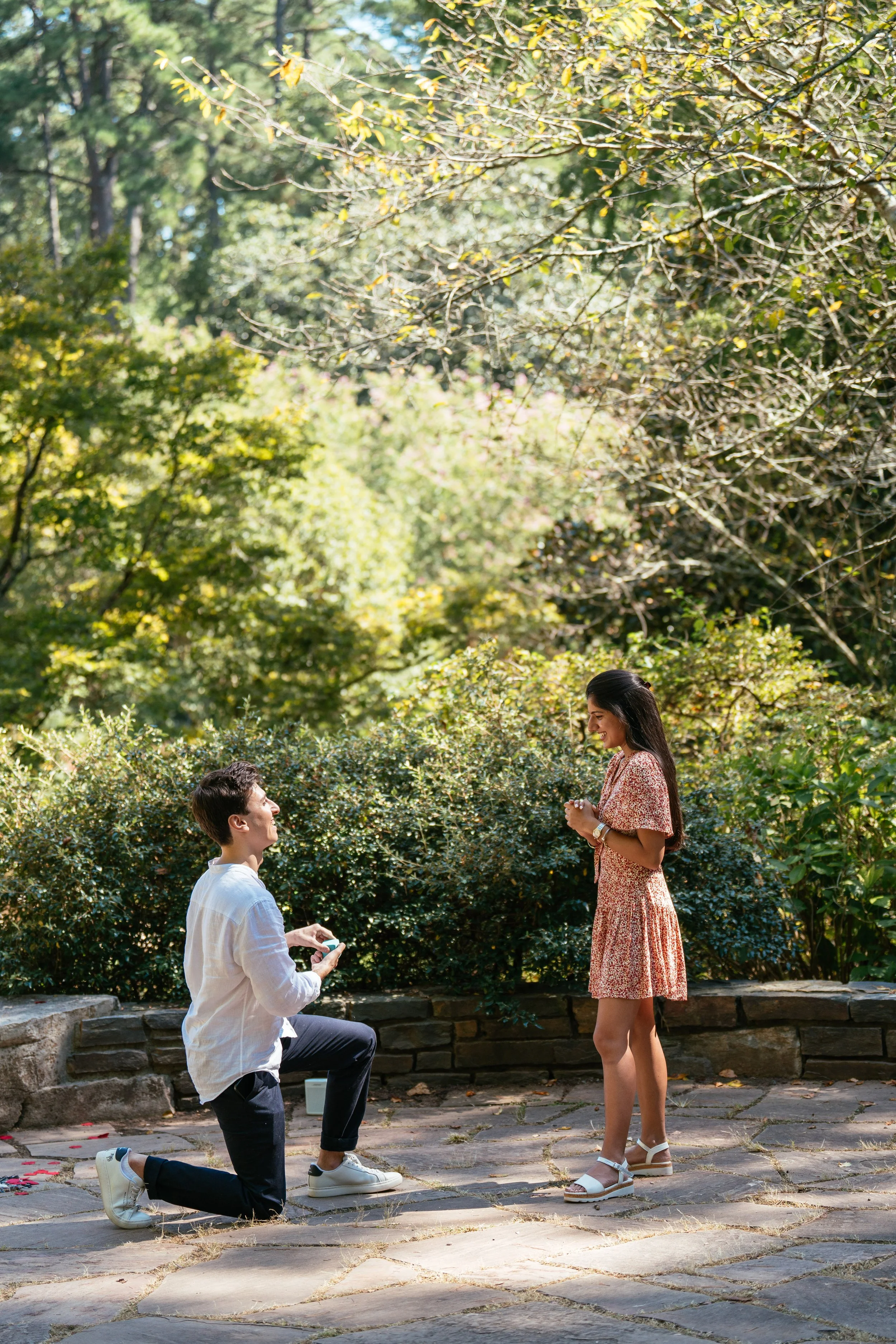 A heartfelt surprise proposal at Duke Gardens in Durham, North Carolina, photographed along a quiet stone pathway surrounded by lush greenery. One partner kneels during the proposal while the other reacts with emotion, capturing the moment naturally 