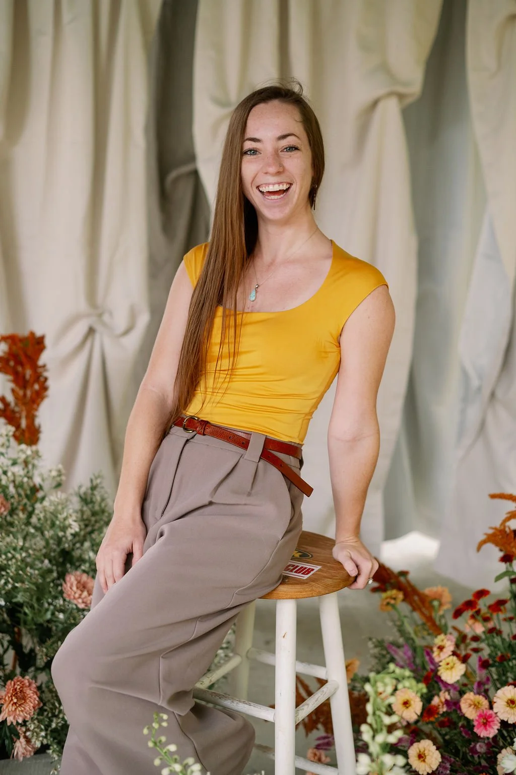 A smiling woman with long brown hair, wearing a yellow sleeveless top and beige high-waisted trousers, sitting on a wooden stool surrounded by flowers and light-colored drapery in the background.