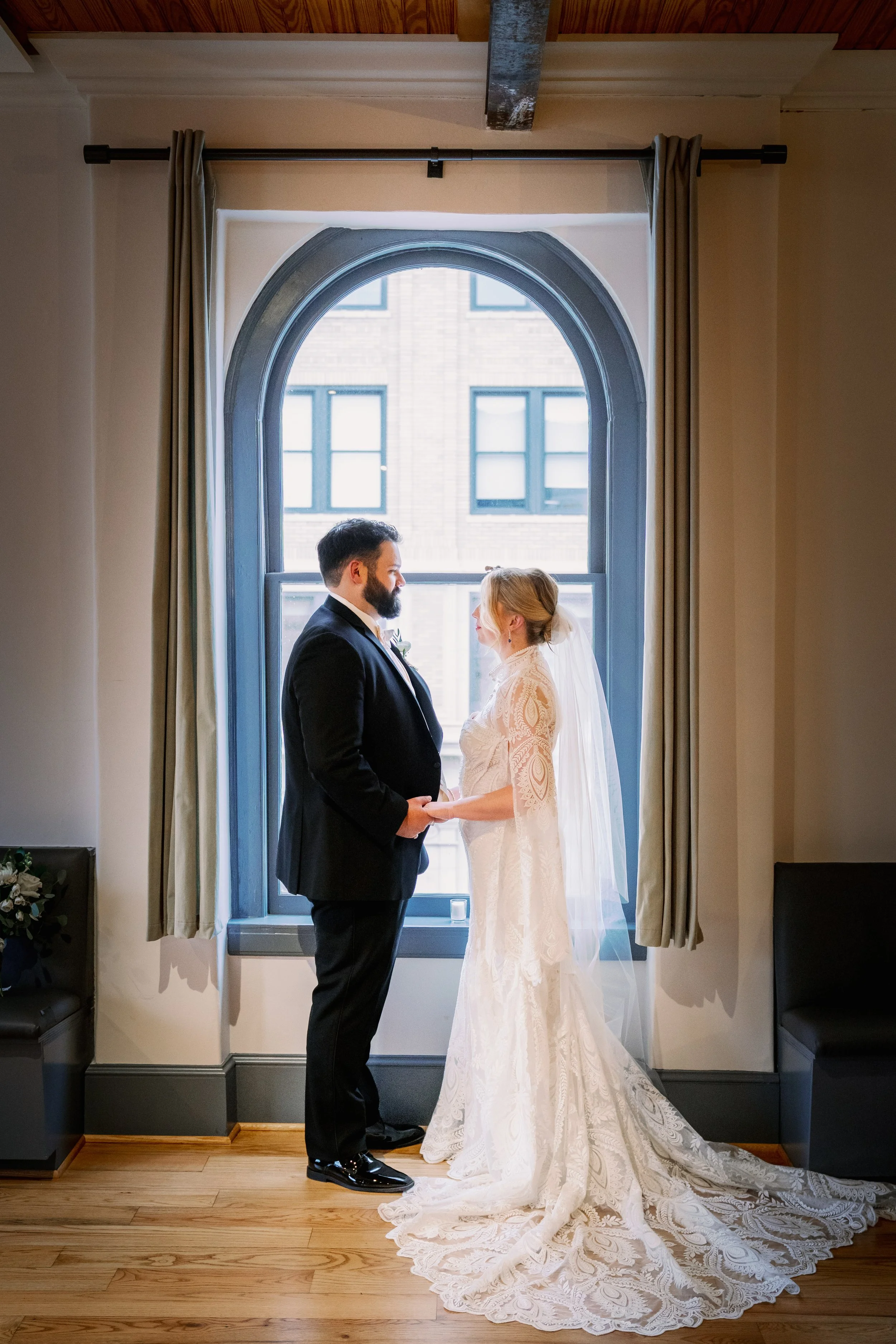 A bride and groom holding hands and looking at each other, standing in front of a large arched window with beige curtains, in a room with wooden flooring and a wooden ceiling beam.