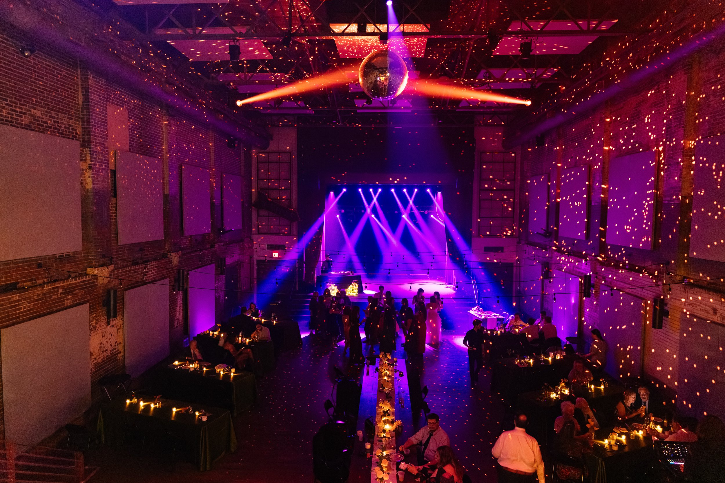 Wide view of a wedding reception at Haw River Ballroom in Saxapahaw North Carolina with colorful stage lighting, disco ball reflections, candlelit tables, and guests dancing inside the historic brick venue