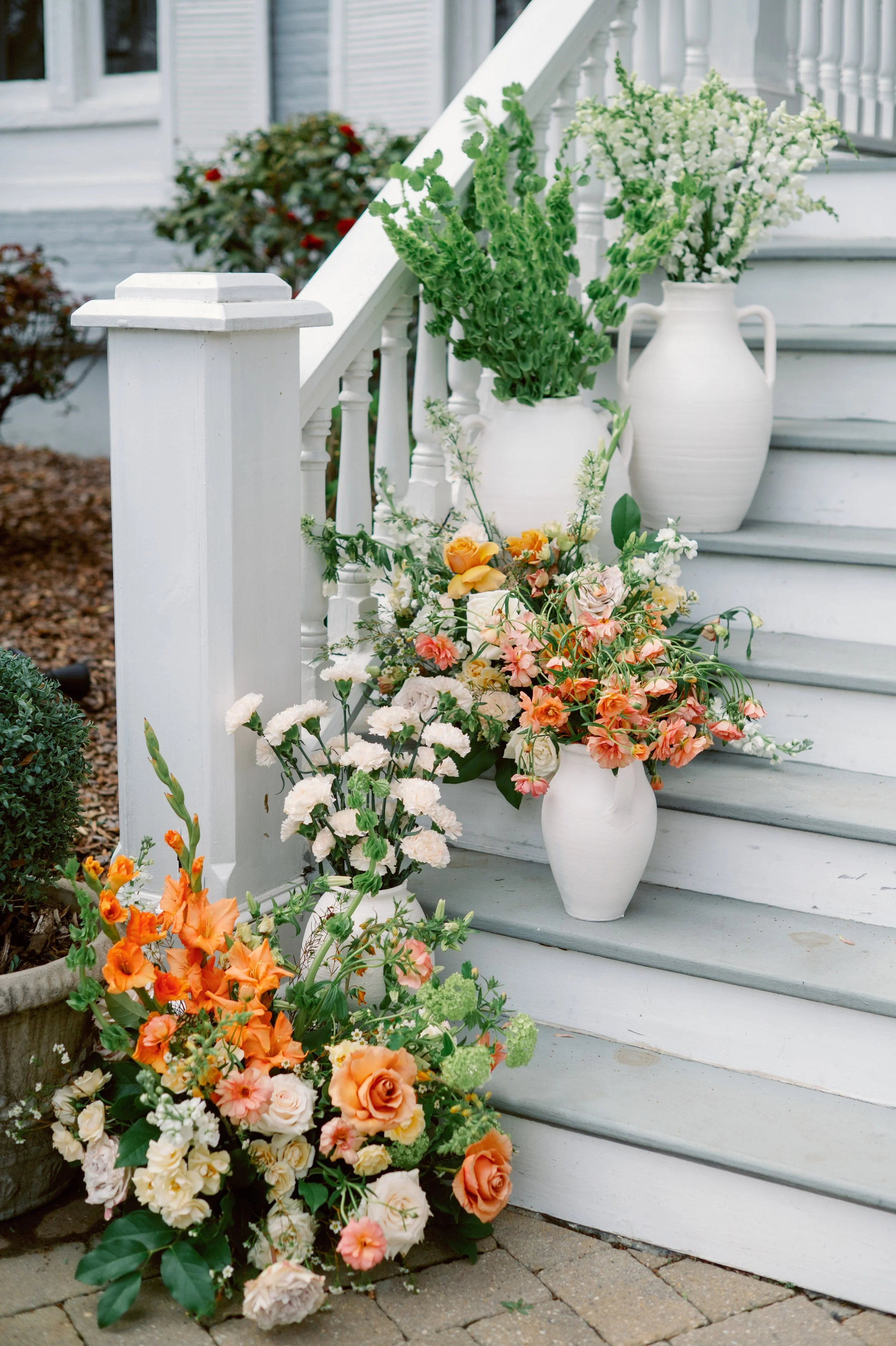 Colorful flower arrangements in white vases on a porch staircase with white railing and stairs, surrounded by potted green plants.