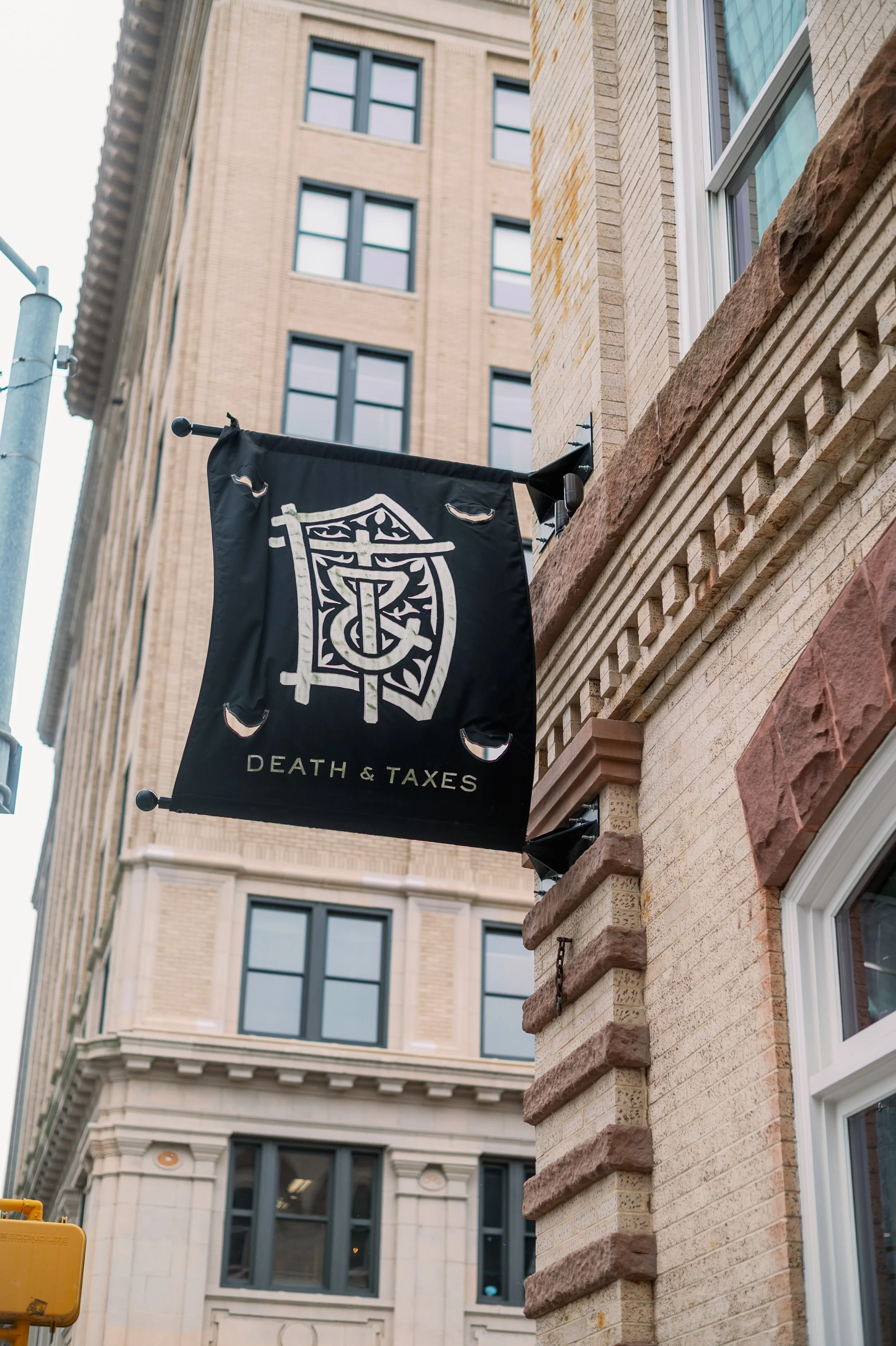 A black outdoor sign with white graphics and text reading 'DEATH & TAXES' is mounted on the brick wall of a building. The sign features a stylized graphic of a horned helmet above the text.