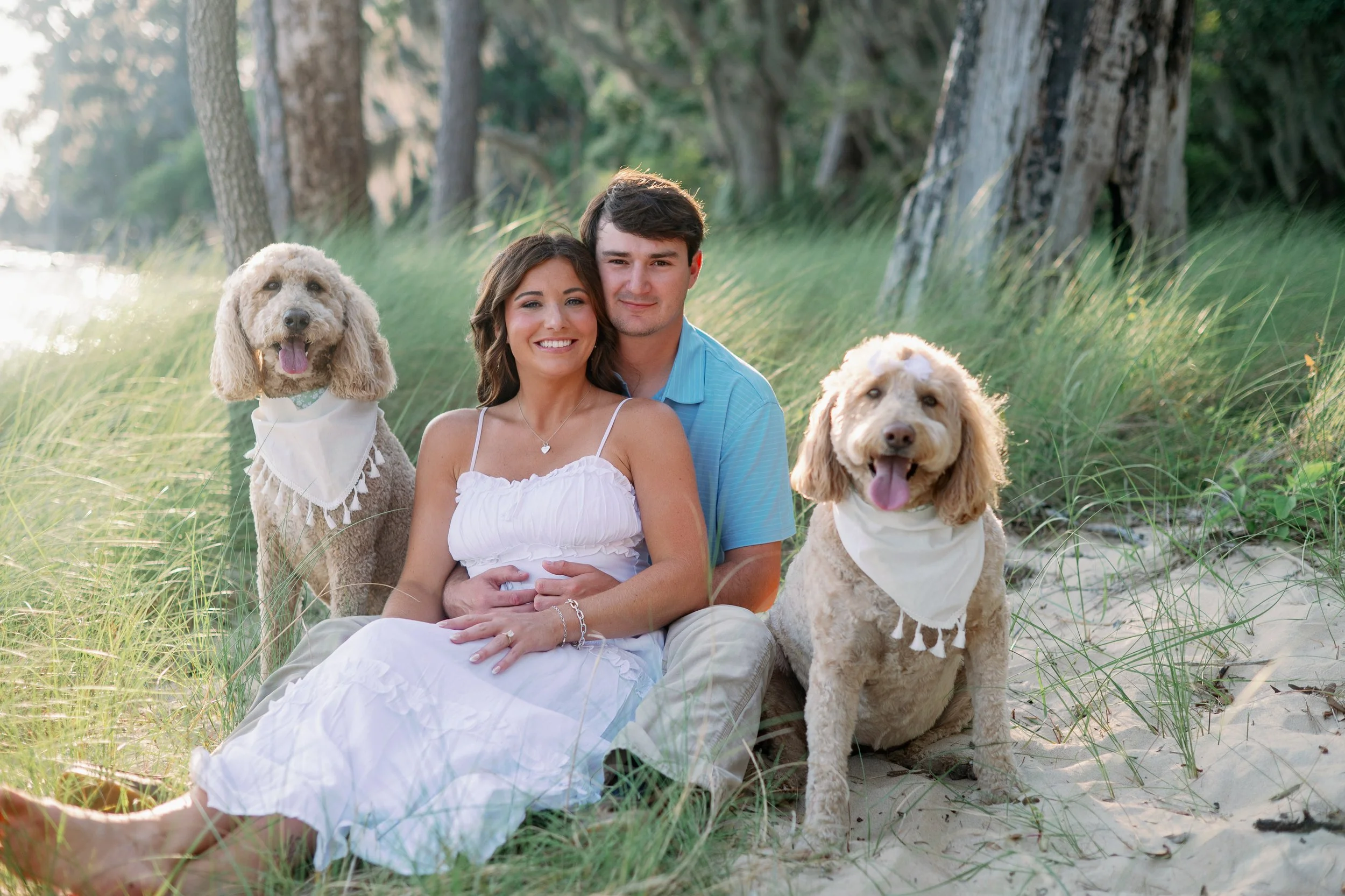 Couple sitting in the sand dunes at Goose Creek State Park Washington NC with their two goldendoodles wearing matching bandanas
