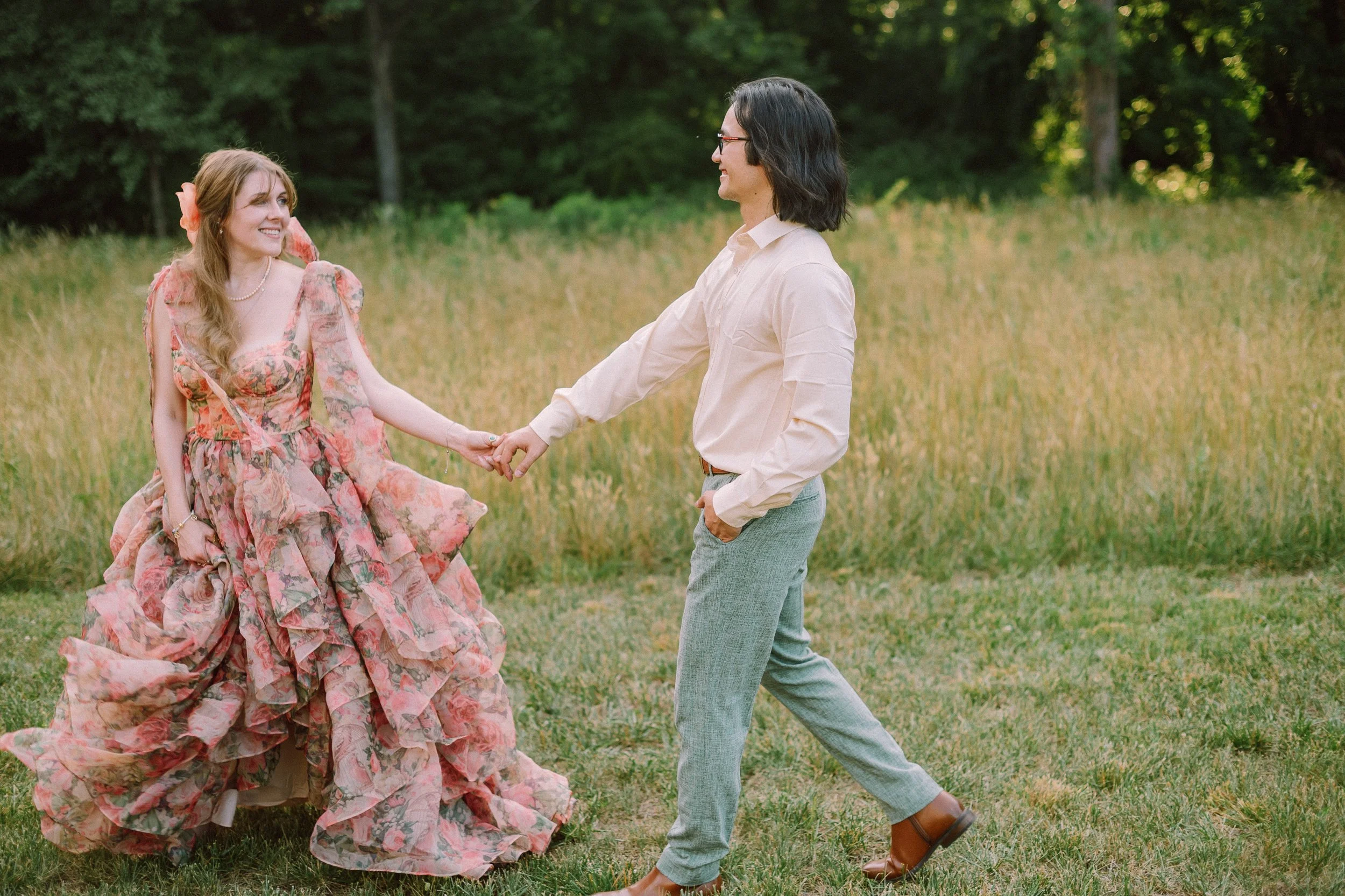 Bride in a pink floral gown holds hands with her groom as they walk through a golden hour field at their intimate family property wedding in Mocksville, North Carolina.