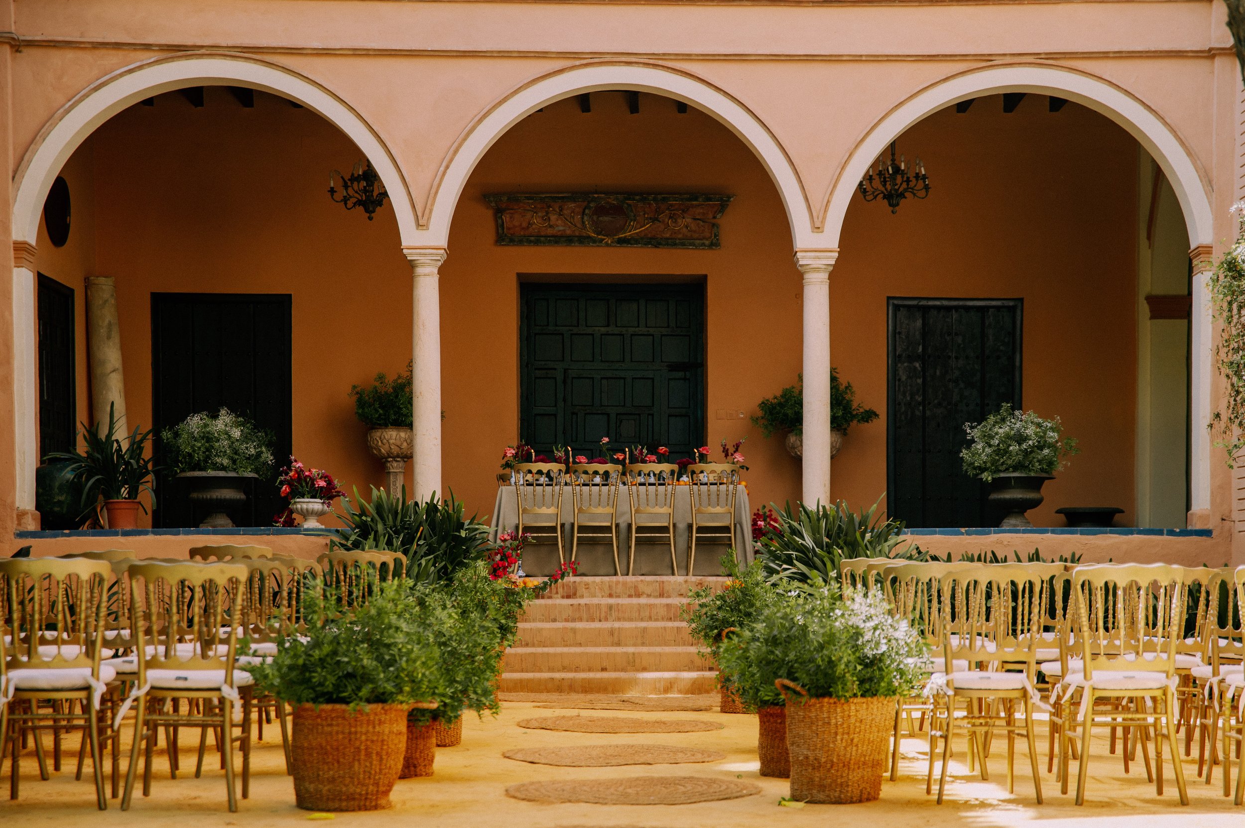 Outdoor decorated event space with beige chairs arranged in rows, potted plants, and a table with chairs on a tiled patio under arches.