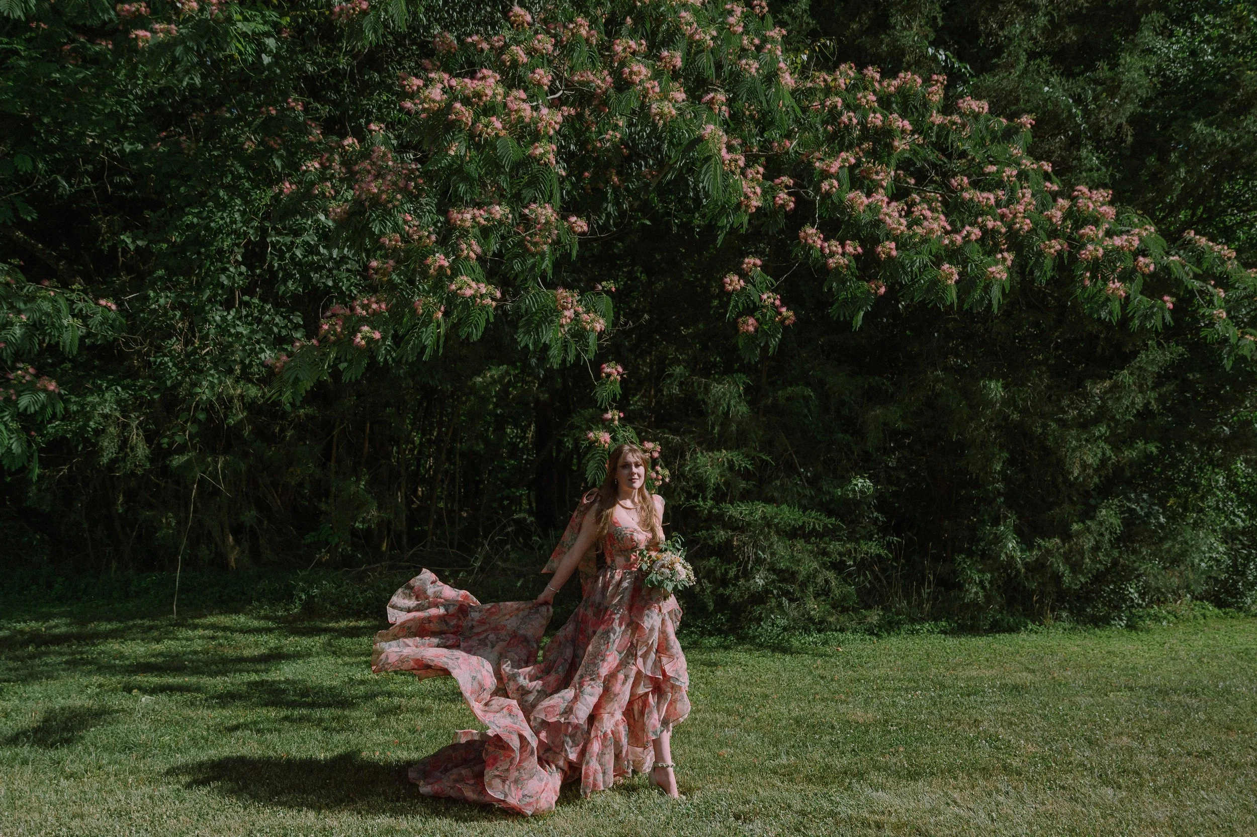 Bride in a flowing pink floral gown tossing her train under a large blooming mimosa tree at a North Carolina family property wedding.