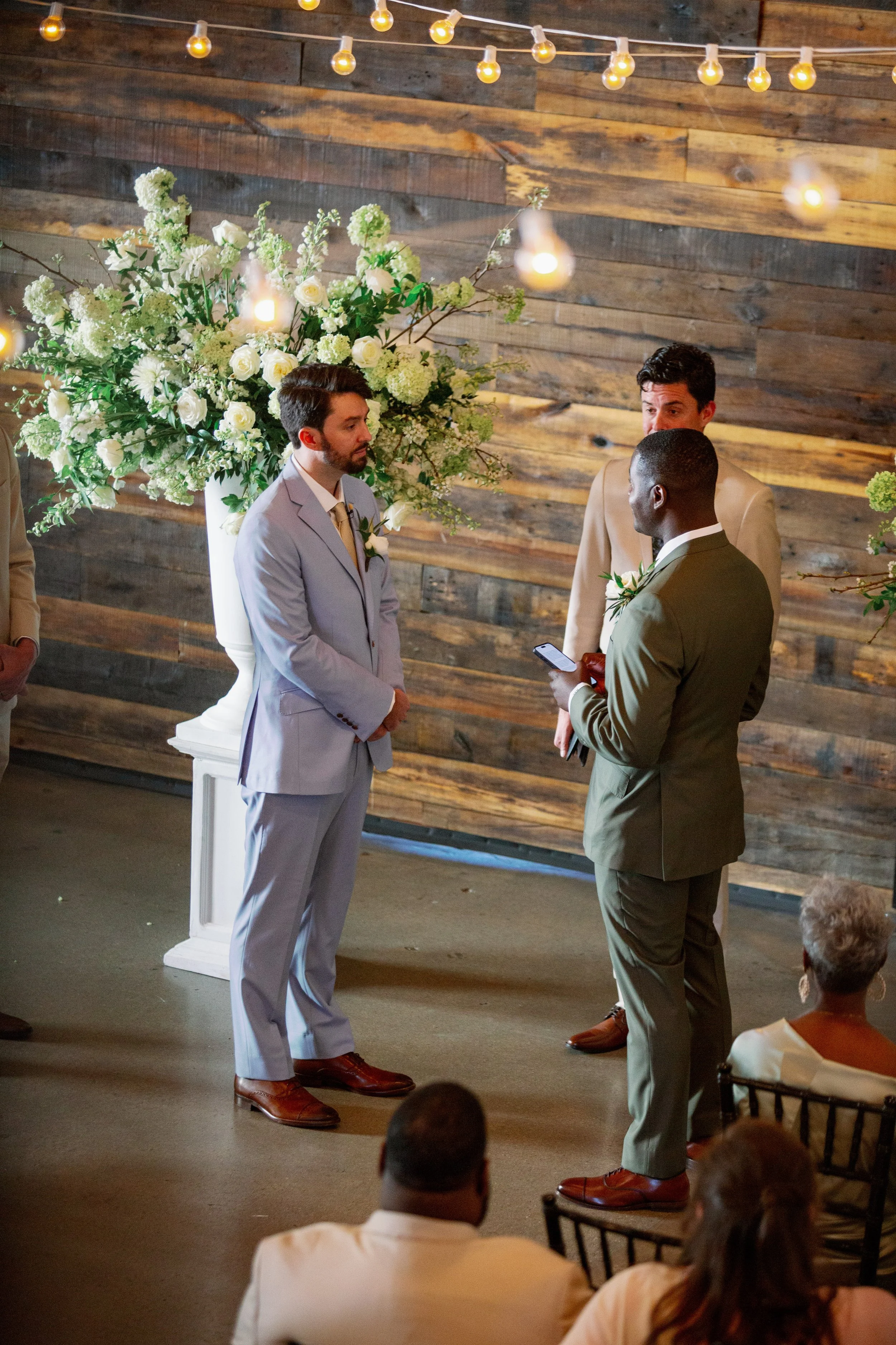 Two grooms stand face to face during their wedding ceremony in front of a reclaimed wood accent wall adorned with string lights and a lush white and green floral arrangement, officiated by their celebrant in Raleigh, NC.