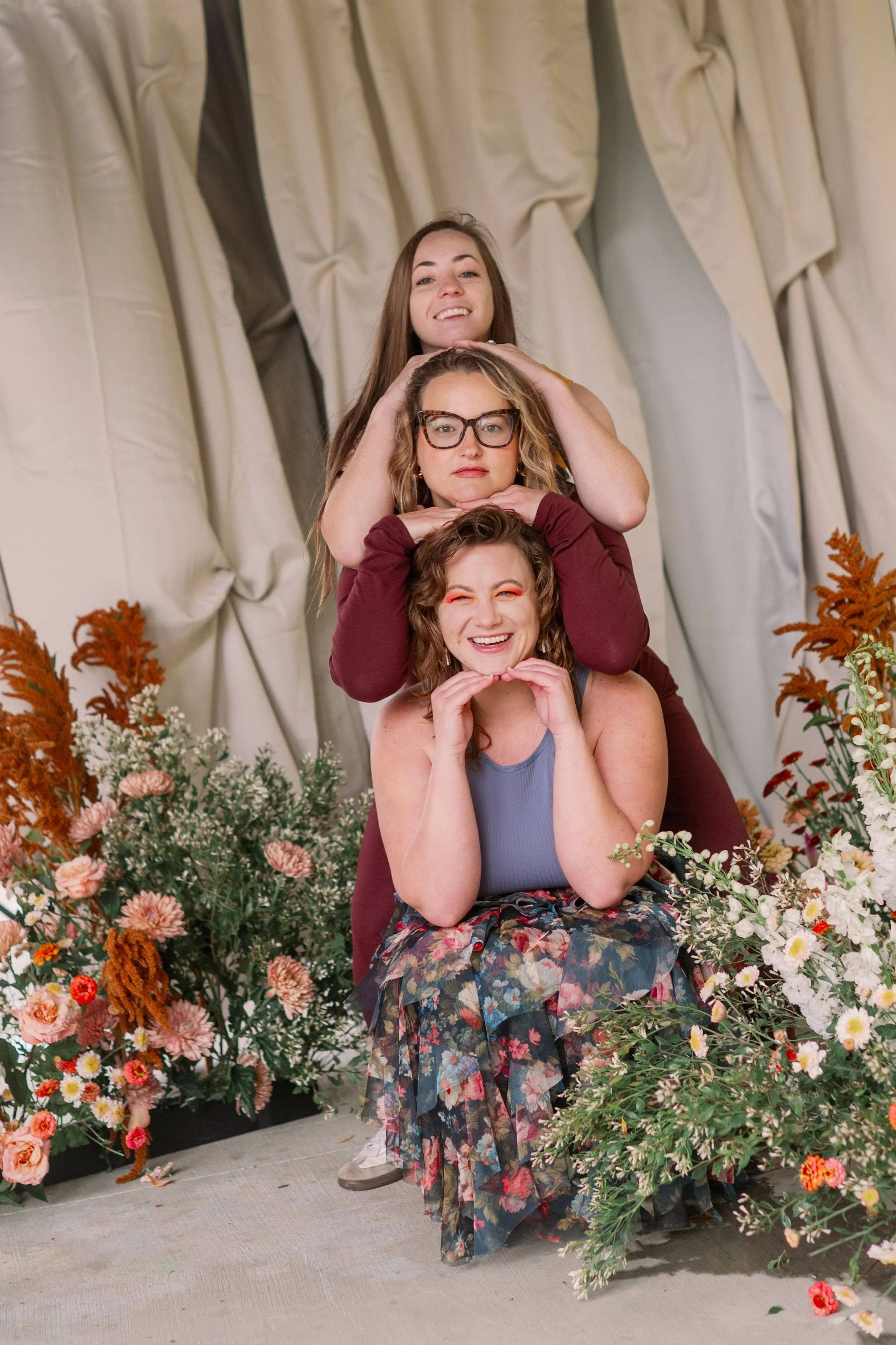 three women stacked in a pose with flowers and greenery around them, smiling and making playful expressions.