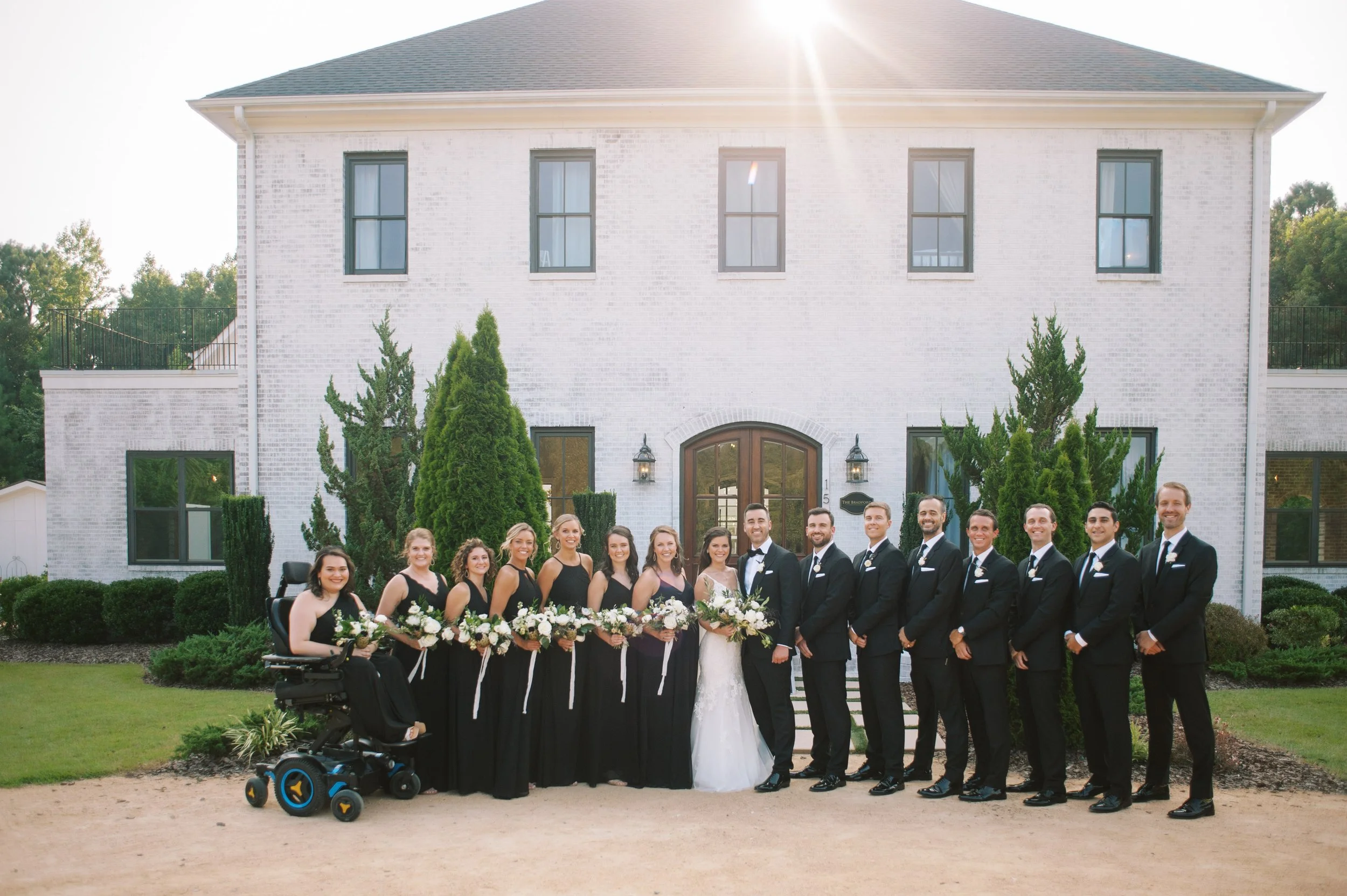 Wedding party standing in front of The Bradford wedding venue in Raleigh, North Carolina, with the bride and groom centered and bridesmaids and groomsmen arranged on the lawn.