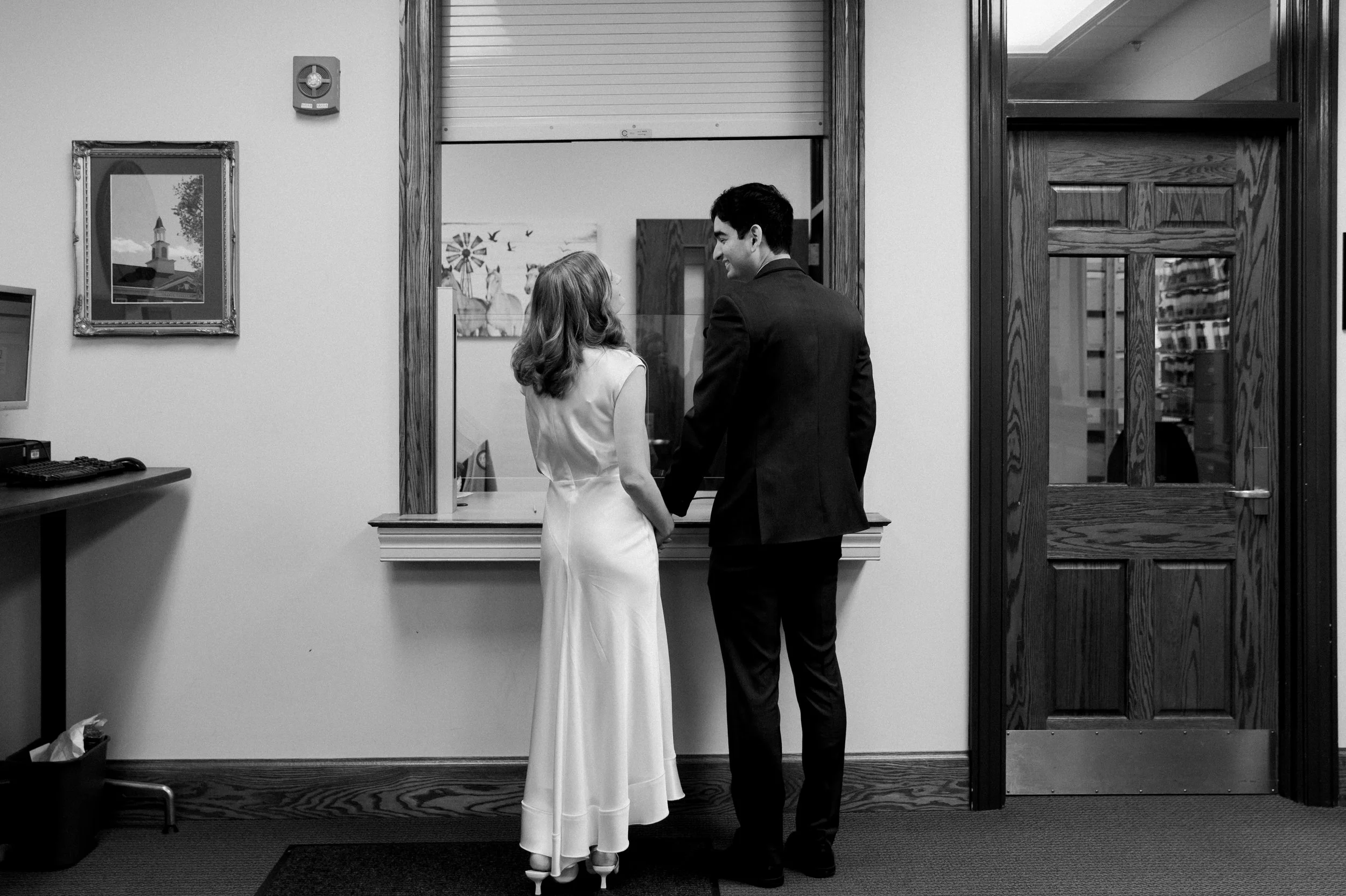 A black and white photo of a man and woman holding hands, standing at a window counter in an office or reception area. The woman wears a white dress and the man is in a dark suit. There is a framed picture on the wall, a computer with a keyboard on a desk, and a doorway with a glass window leading to another room.