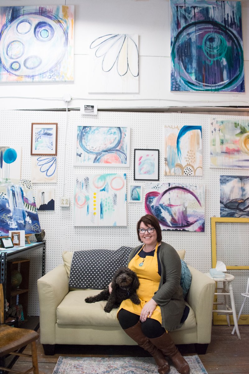  Jaci sitting in her studio with her dog Ellie. Behind them is a wall full of abstract paintings.&nbsp; 