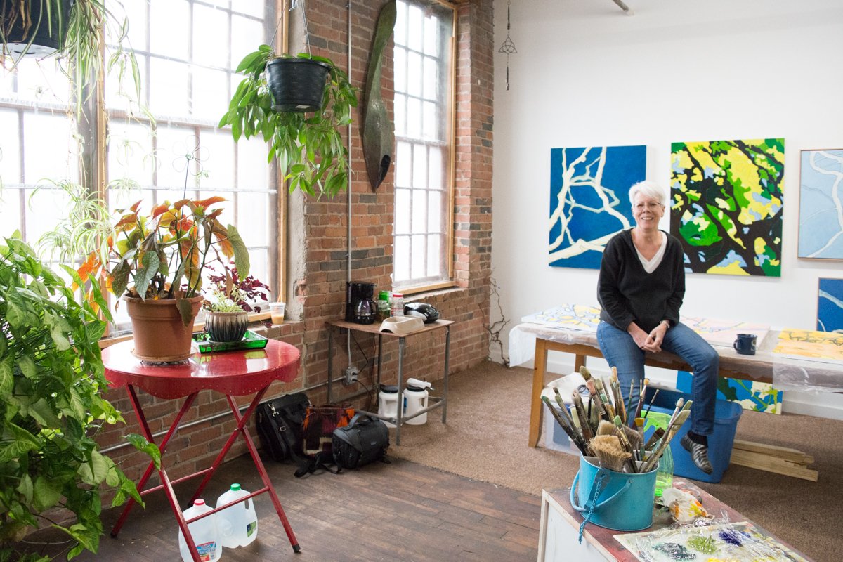  Steph sitting on her studio table surrounded by paintings and plants.&nbsp; 