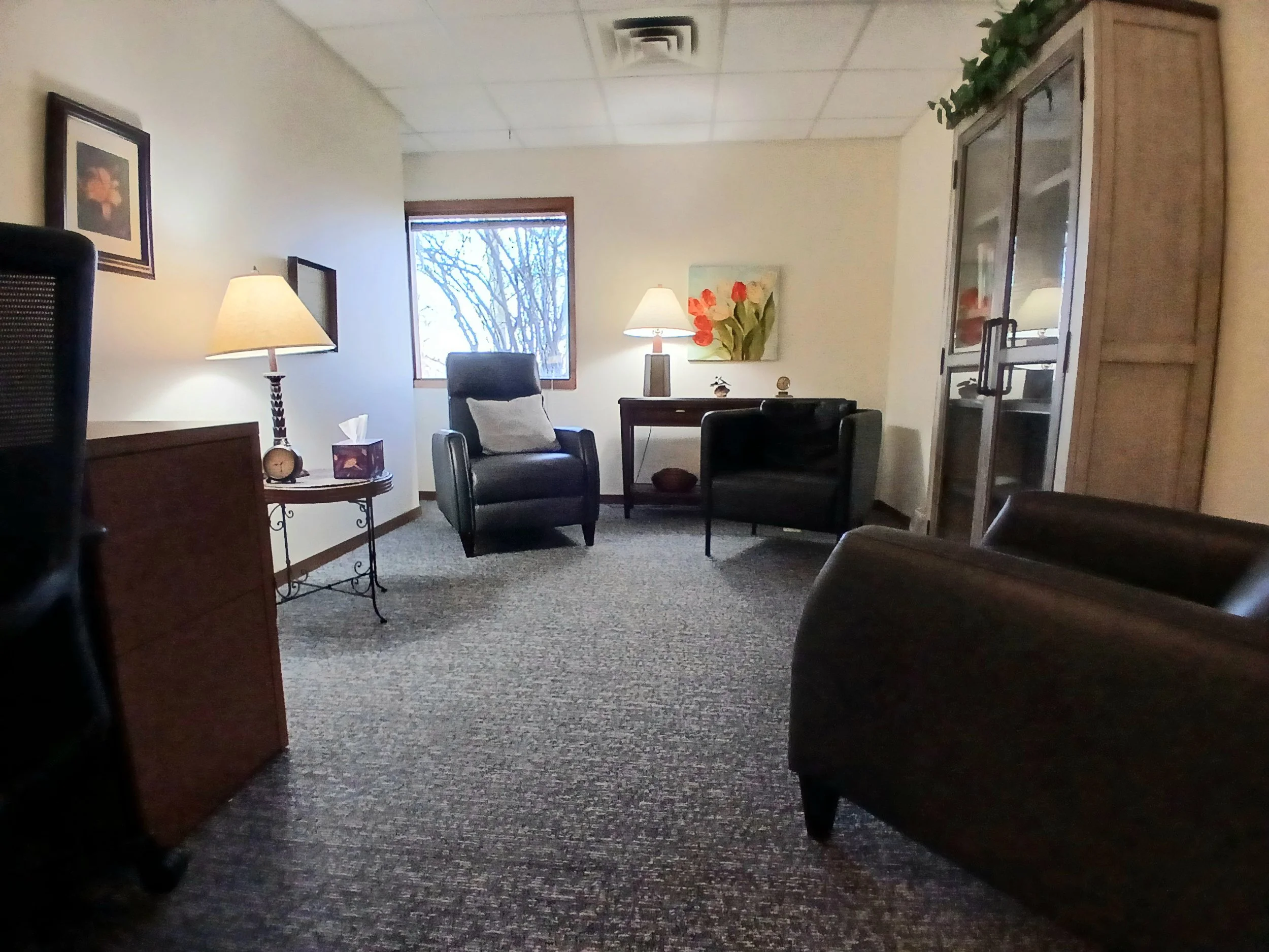 An office with two black armchairs, a wooden side table with a lamp, a tissue box, and a decorative clock. There is a window showing trees outside, a painting of red flowers on the wall, and a glass-fronted cabinet.