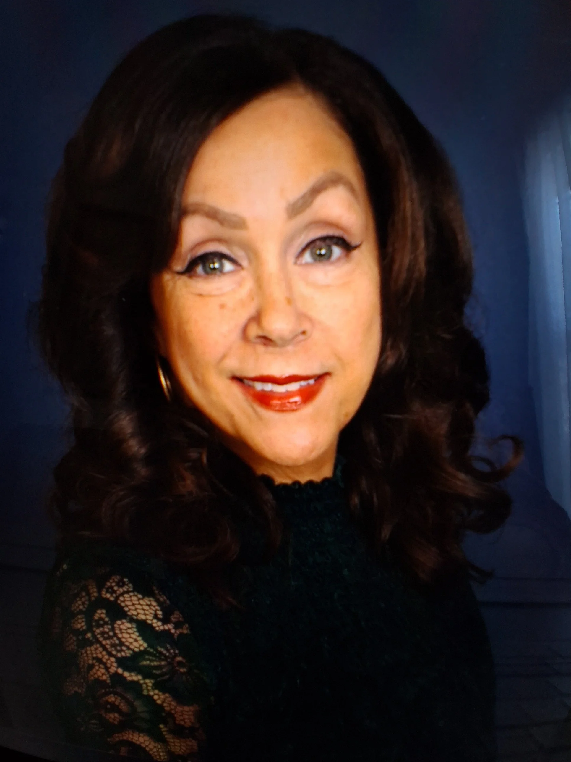 A woman with dark, curly hair, fair skin, wearing makeup including red lipstick and eye makeup, dressed in a black lace top, smiling at the camera against a dark background.