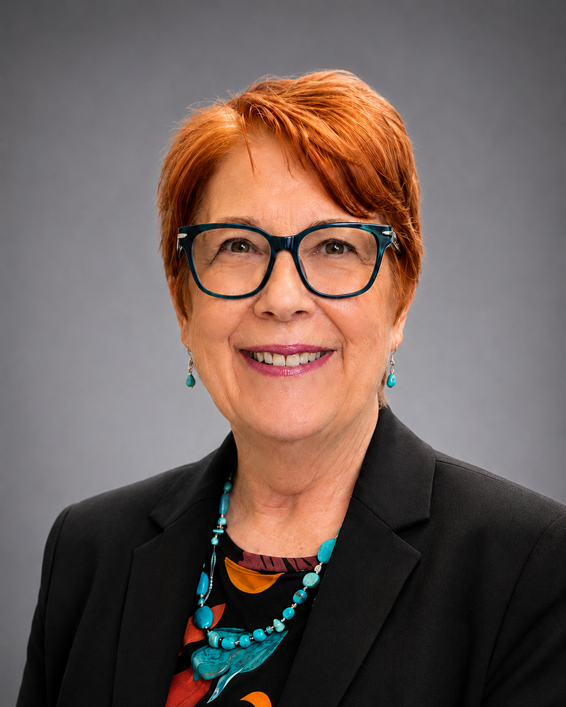 Portrait of a smiling woman with short red hair and wearing large glasses, turquoise jewelry, and a black blazer against a gray background.