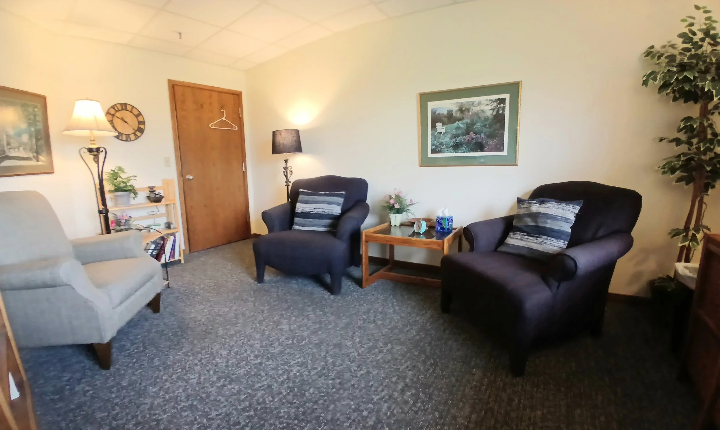 A cozy living room with two dark armchairs, a light-colored sofa, a small wooden table with a tissue box and flowers, a bookshelf, and wall art. There are two standing lamps and a large tall artificial plant.