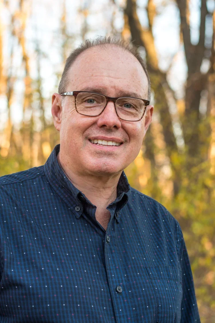 A smiling man with glasses and receding hair standing outdoors with trees in the background.