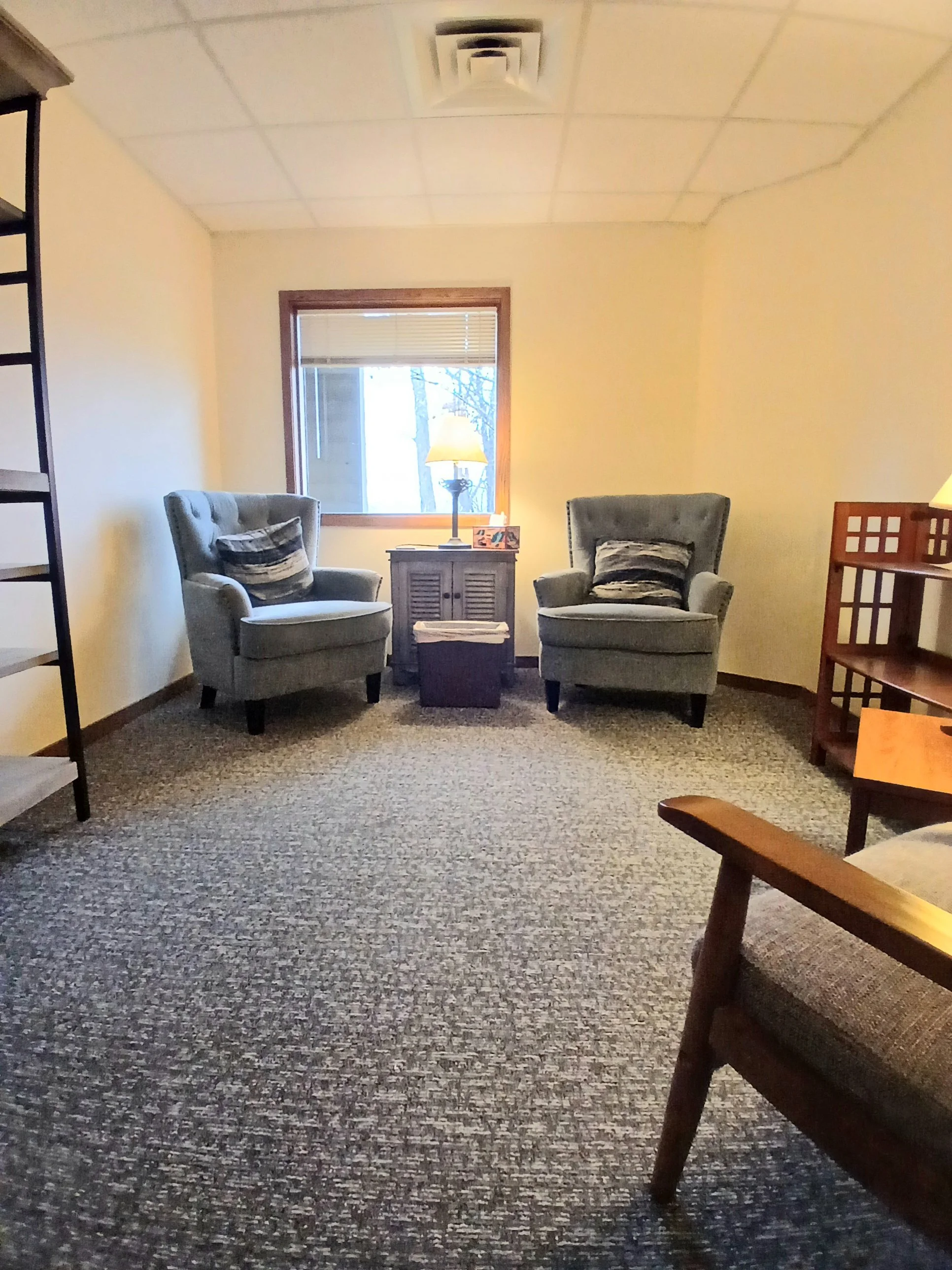 A cozy living room with two armchairs, a small wooden table with a lamp on it, a window with a blind, and a wooden stair railing. The carpet has a speckled pattern, and there are pillows on the armchairs.