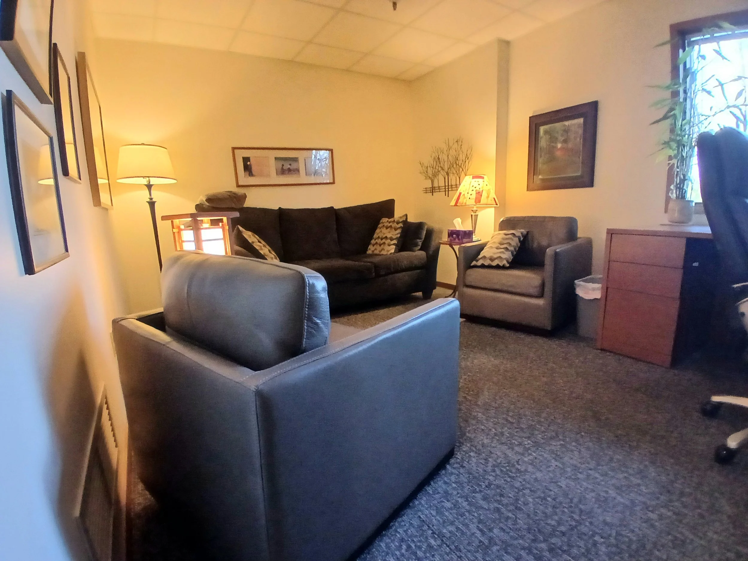 A cozy therapy office with a beige wall, a framed picture, a tall floor lamp, a dark brown sofa with patterned pillows, a side lamp, a beige armchair, and a window with a plant on the windowsill.