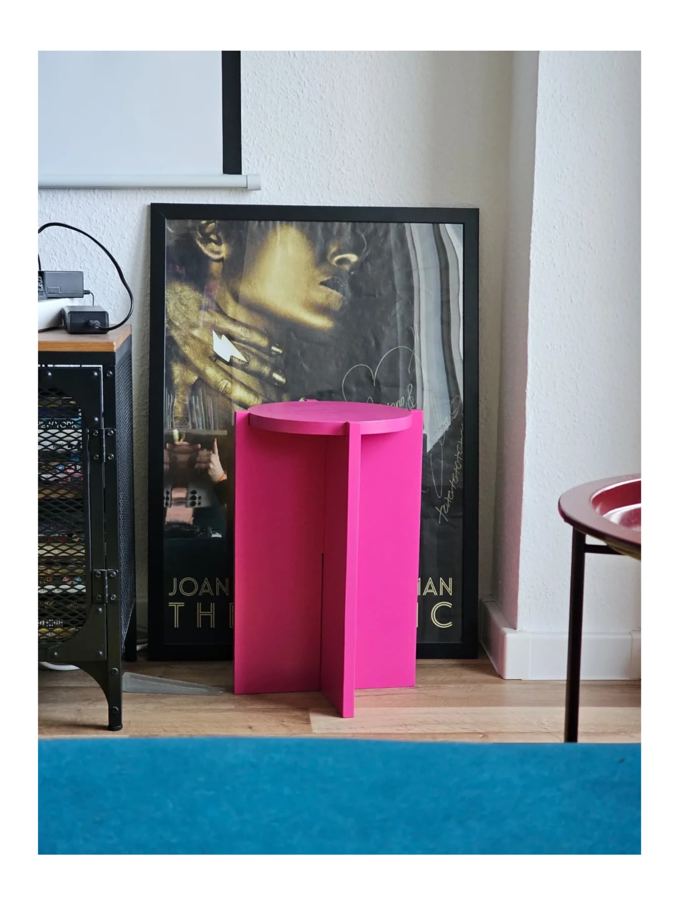 hot pink side table slotted together with round table top, paced in front of an art work on the floor, leaning against the wall