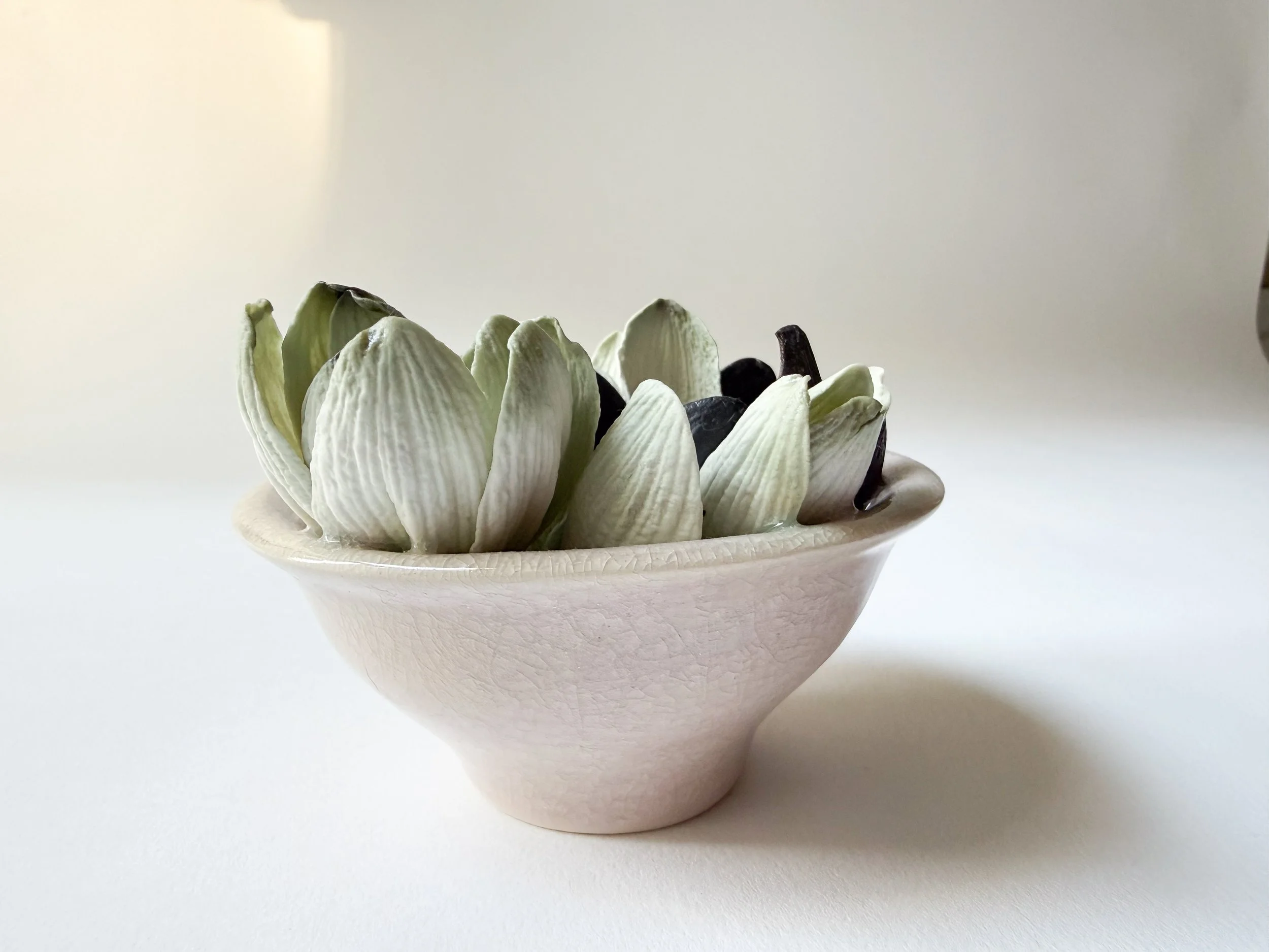 A small, white ceramic bowl with a textured surface filled with white porcelain magnolia petals, set against a plain white background.