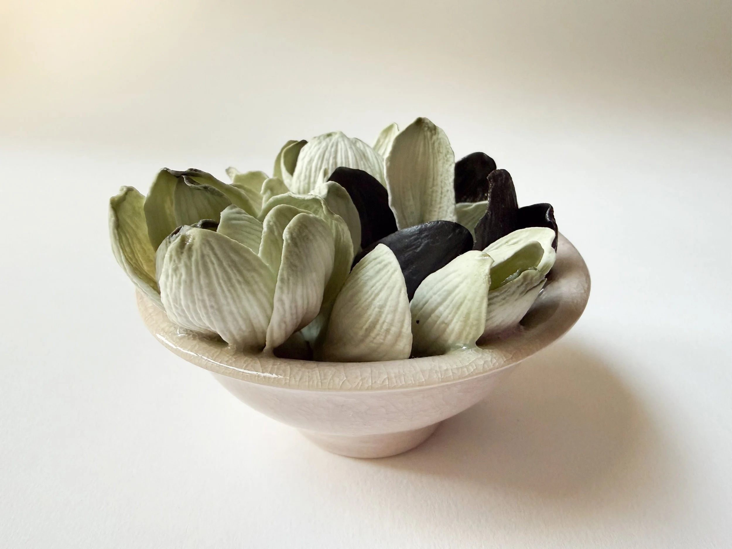 A small ceramic bowl filled with black, white, porcelain magnolia petals on a plain, light-colored surface.