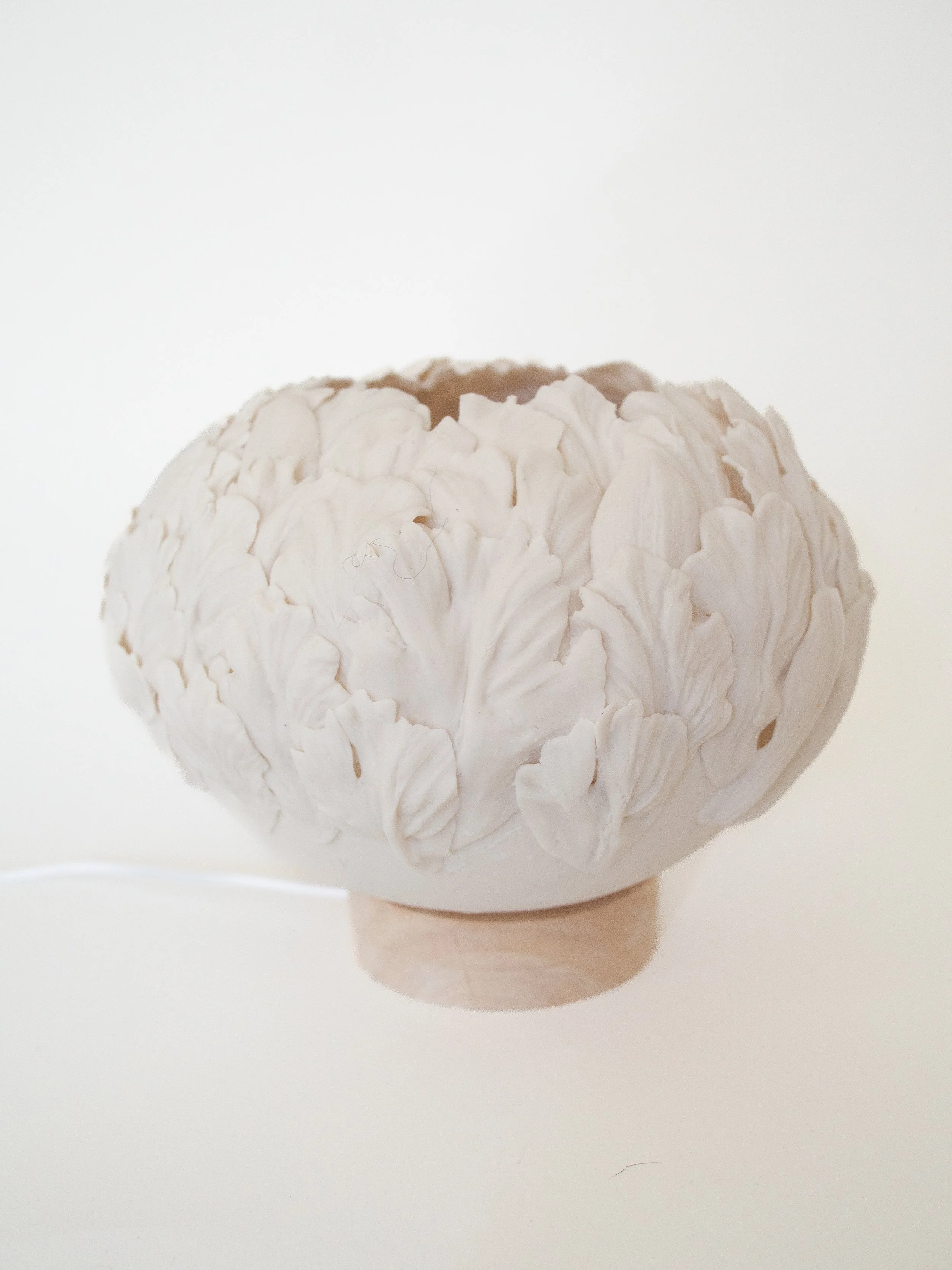 White porcelain ceramic lamp sculpture with intricate folds of flower petals, placed on a small wooden base.