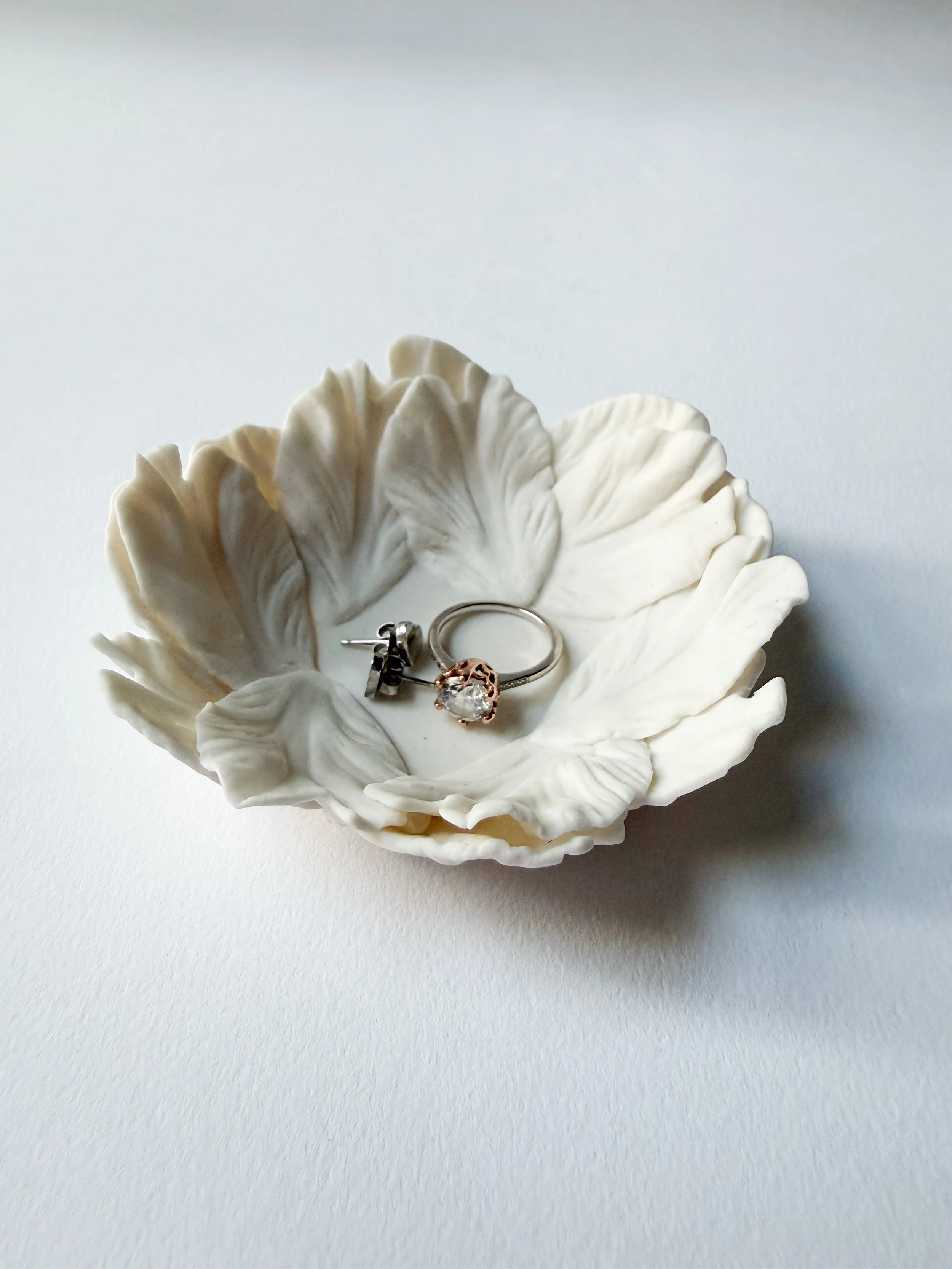 A white porcelain ceramic dish in the shape of a flower with ruffled petals, holding a silver ring with a pink gemstone and a pair of silver earrings with black and clear stones, on a white surface.