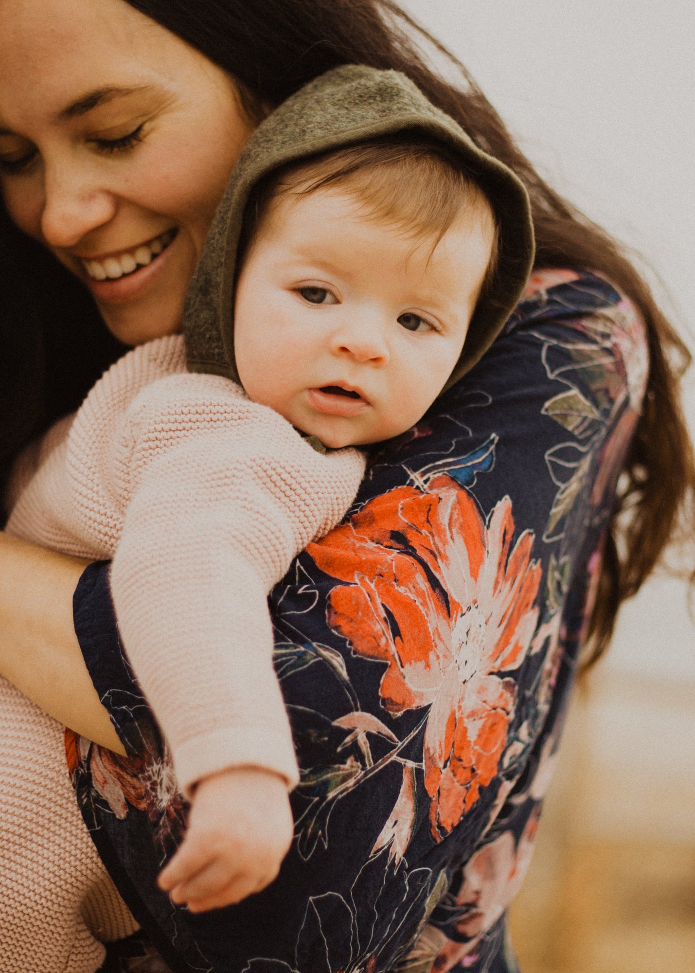 Smiling mother in floral shirt, holding newborn baby in a hooded sweater.