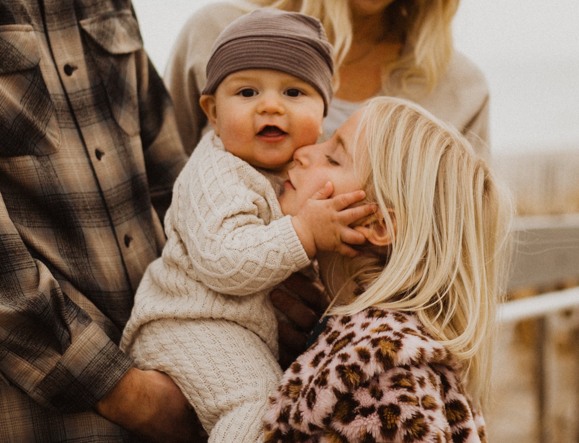 Smiling baby in a woolden hat, looking at camera and touching mother's face while being held up by father.