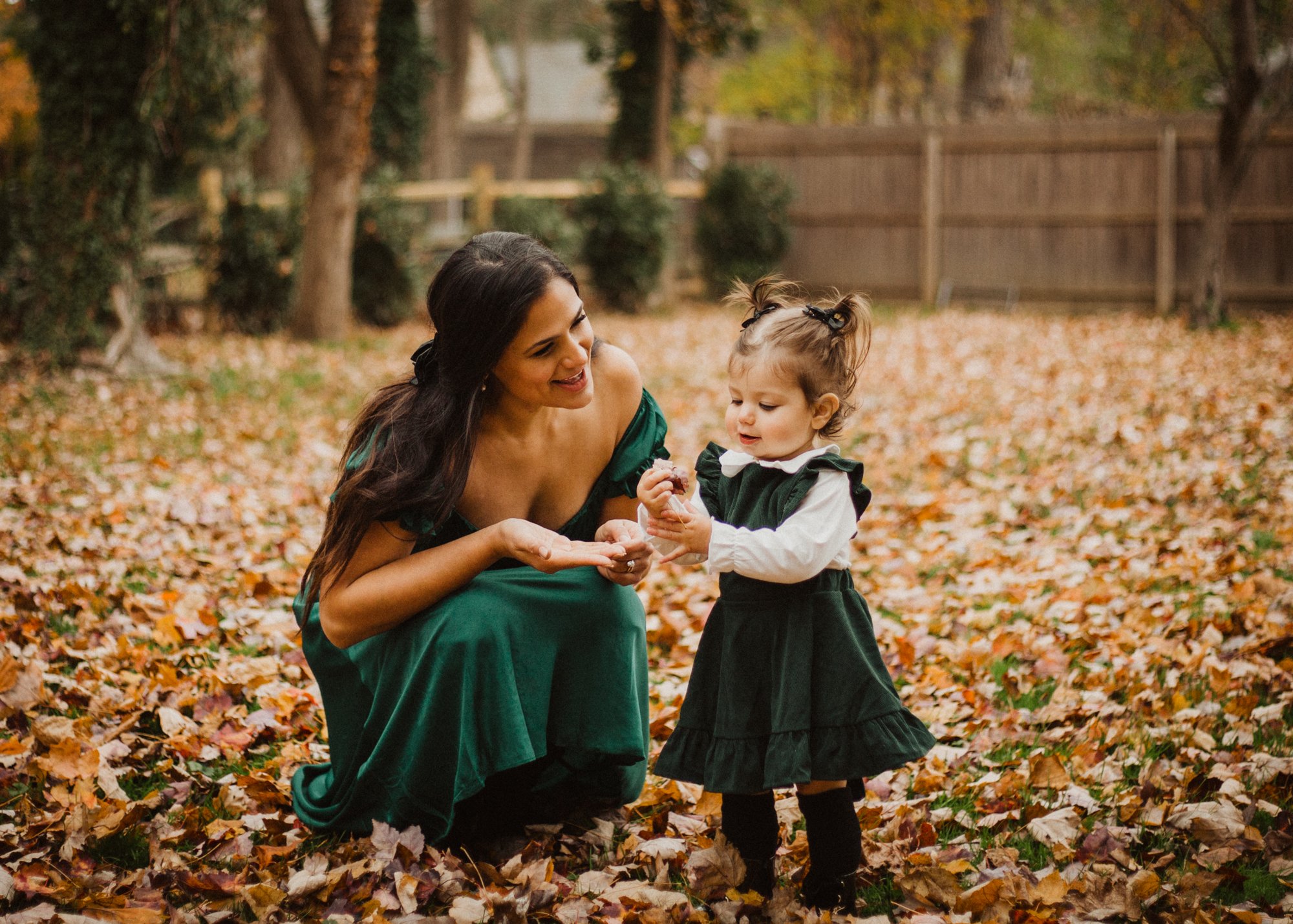 Mother and young daughter in matching green outfits, crouched in fallen autumn leaves.