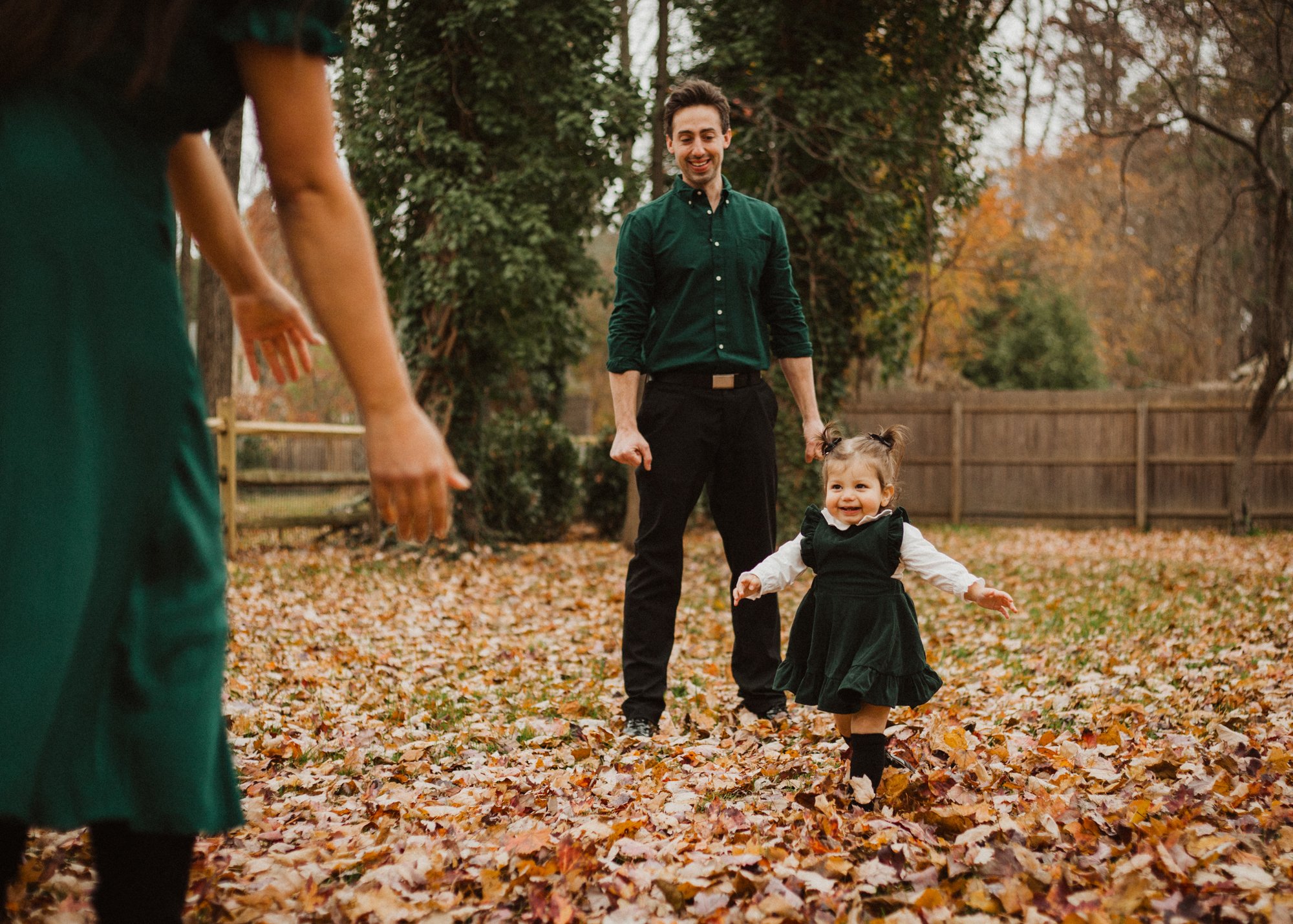 Family in matching green outfits, with young girl smiling and running between parents across fallen leaves.