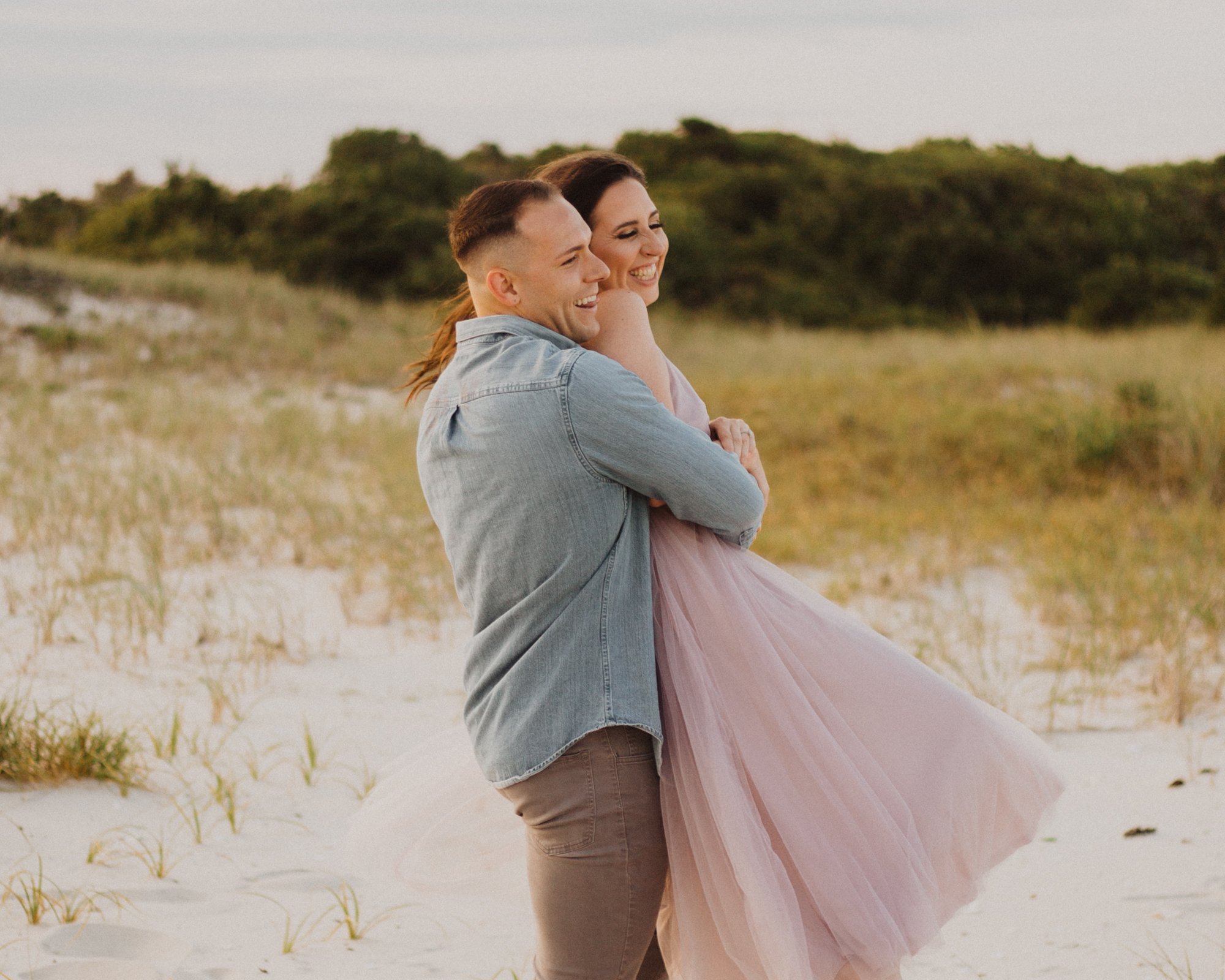 Smiling man in blue shirt holding and lifting his bride from behind as she laughs. In grassy sand dunes.