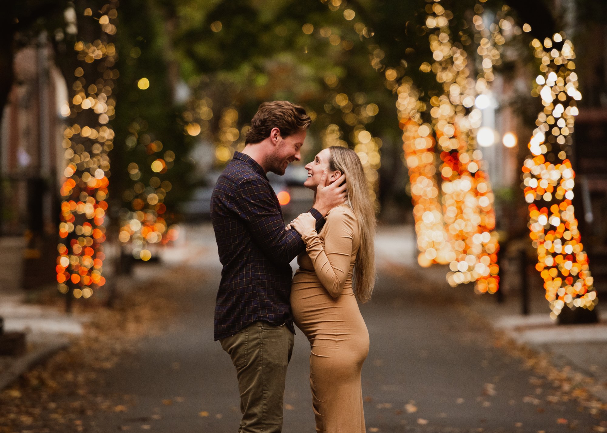 Pregnant woman and husband looking into each others eyes and smiling in the middle of Philadelphia pedestrian walkway. Trees with fairy lights in background.