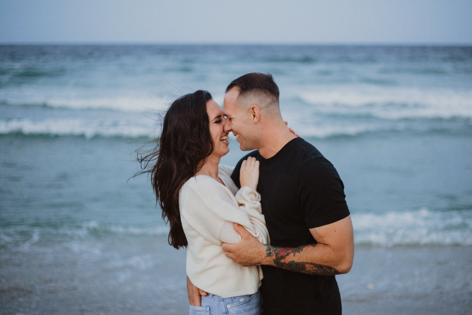 Woman in white shirt and man wearing black, smilling at each other in close embrace on beach with ocean backdrop.