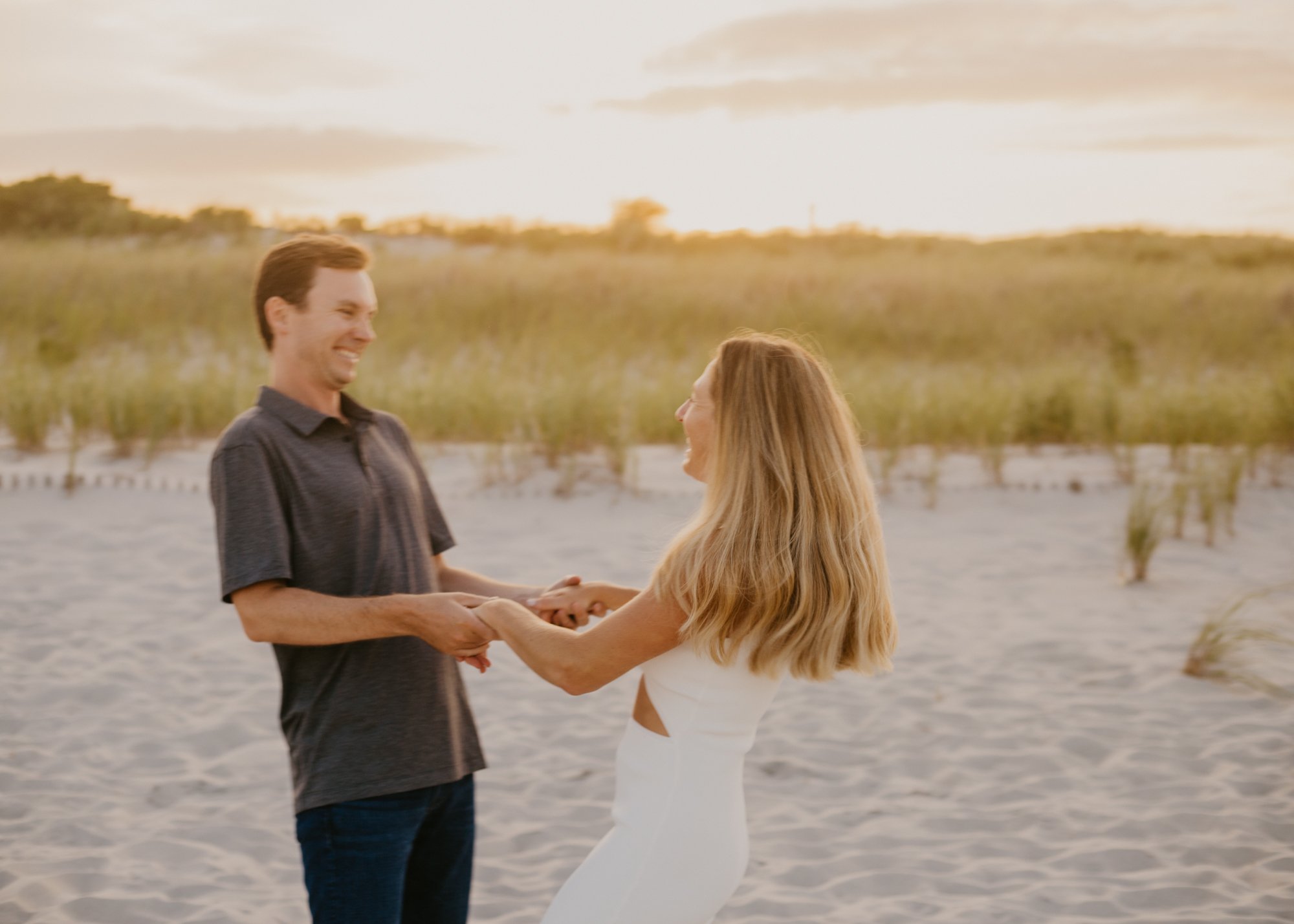 Man smiling at and holding hand with woman, twirling around each other on beach in natural light at sunset.