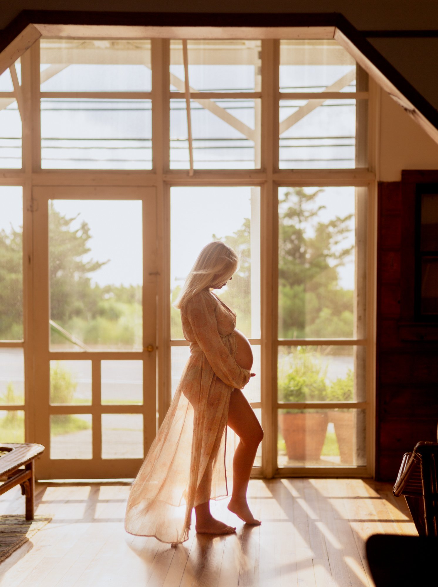 Pregnant woman wearing sheer gown, standing in natural light and cradling her belly, framed in wooden beach house door at the Jersey shore.