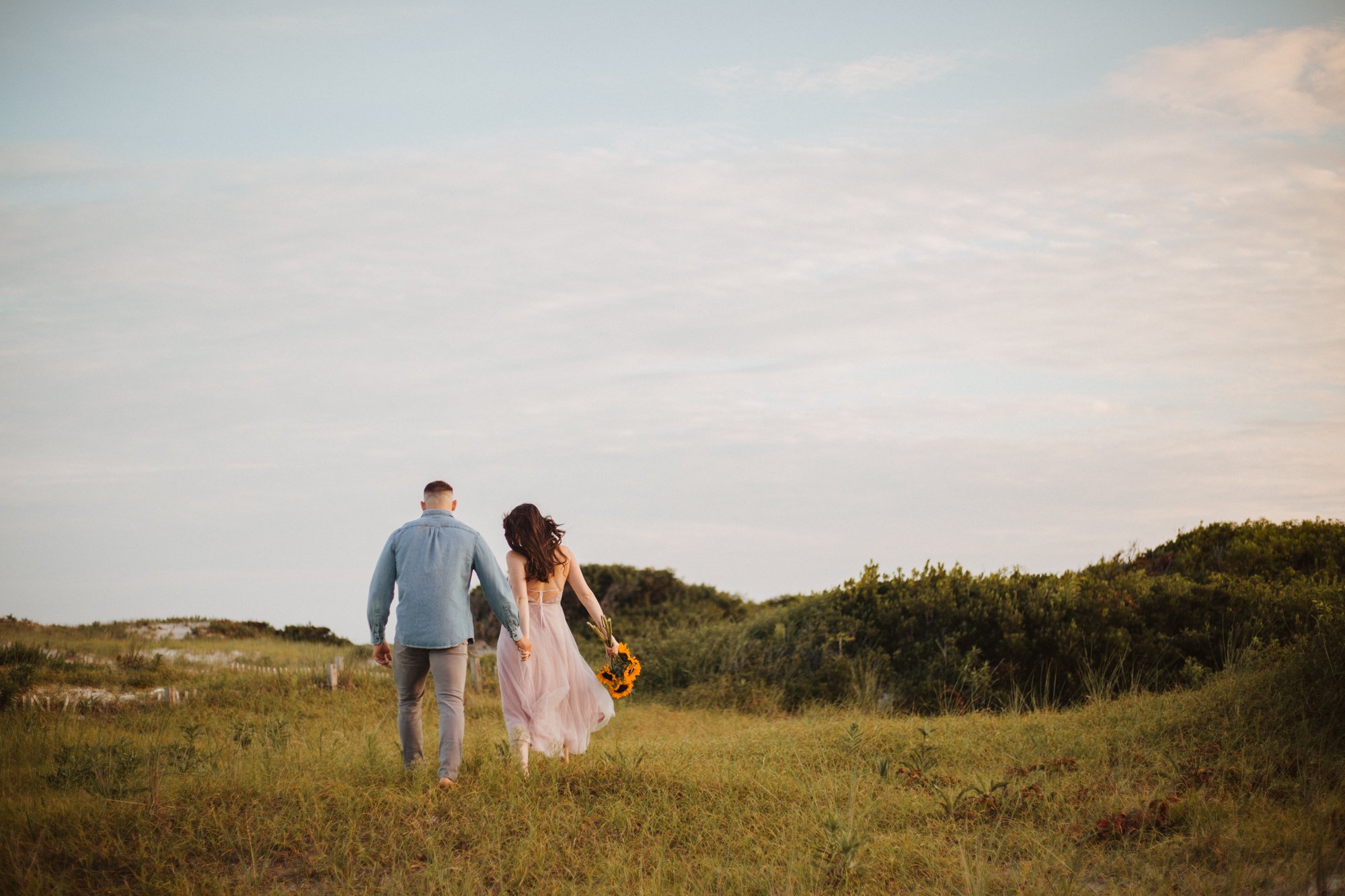 Man in blue shirt and woman in white dress walking through grassy scrub of Jersey shore sand dunes.