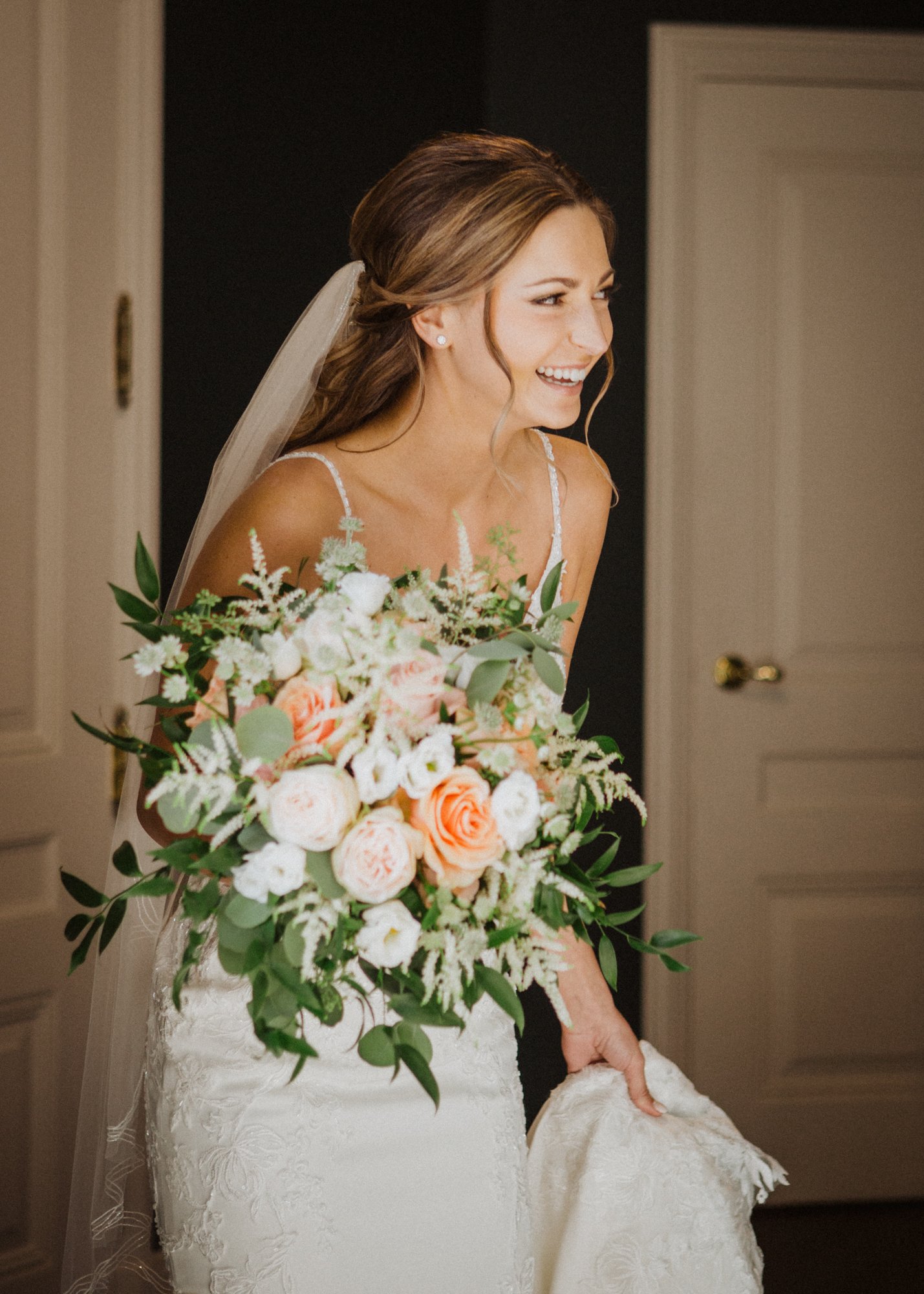 Bride smilling, wearing wedding dress and holding flower bouquet, framed in open doorway.